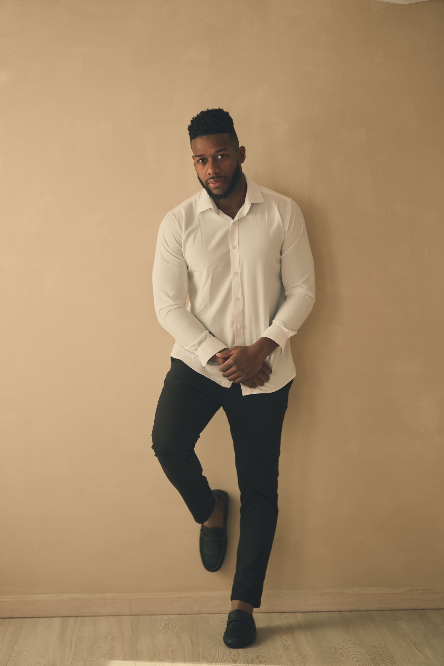 Professional fashion portrait of person in white button-up shirt and dark pants posing against beige wall.
