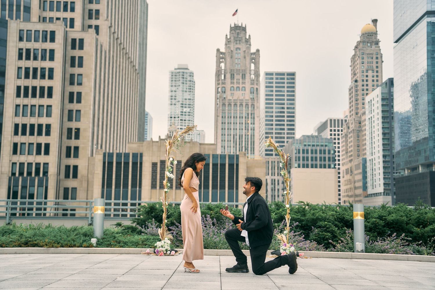 A romantic marriage proposal takes place in front of a cityscape with tall buildings and greenery in the background.