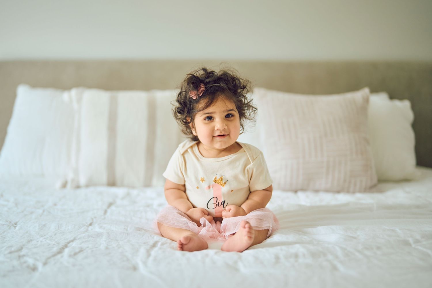 A small child in a beige onesie sits on a white bed with pillows behind them.