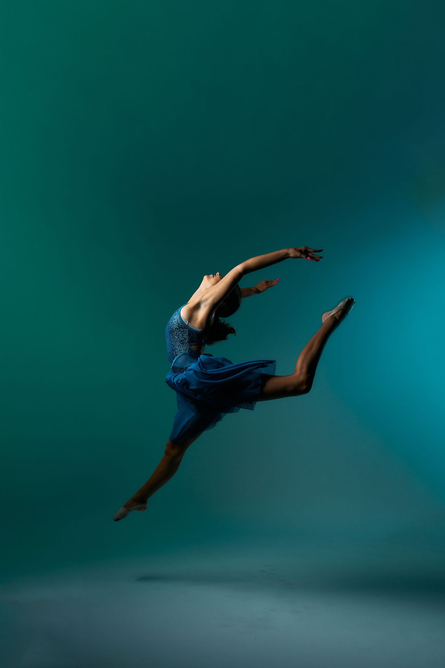 A dancer in blue leaps gracefully against a teal background in dramatic lighting.