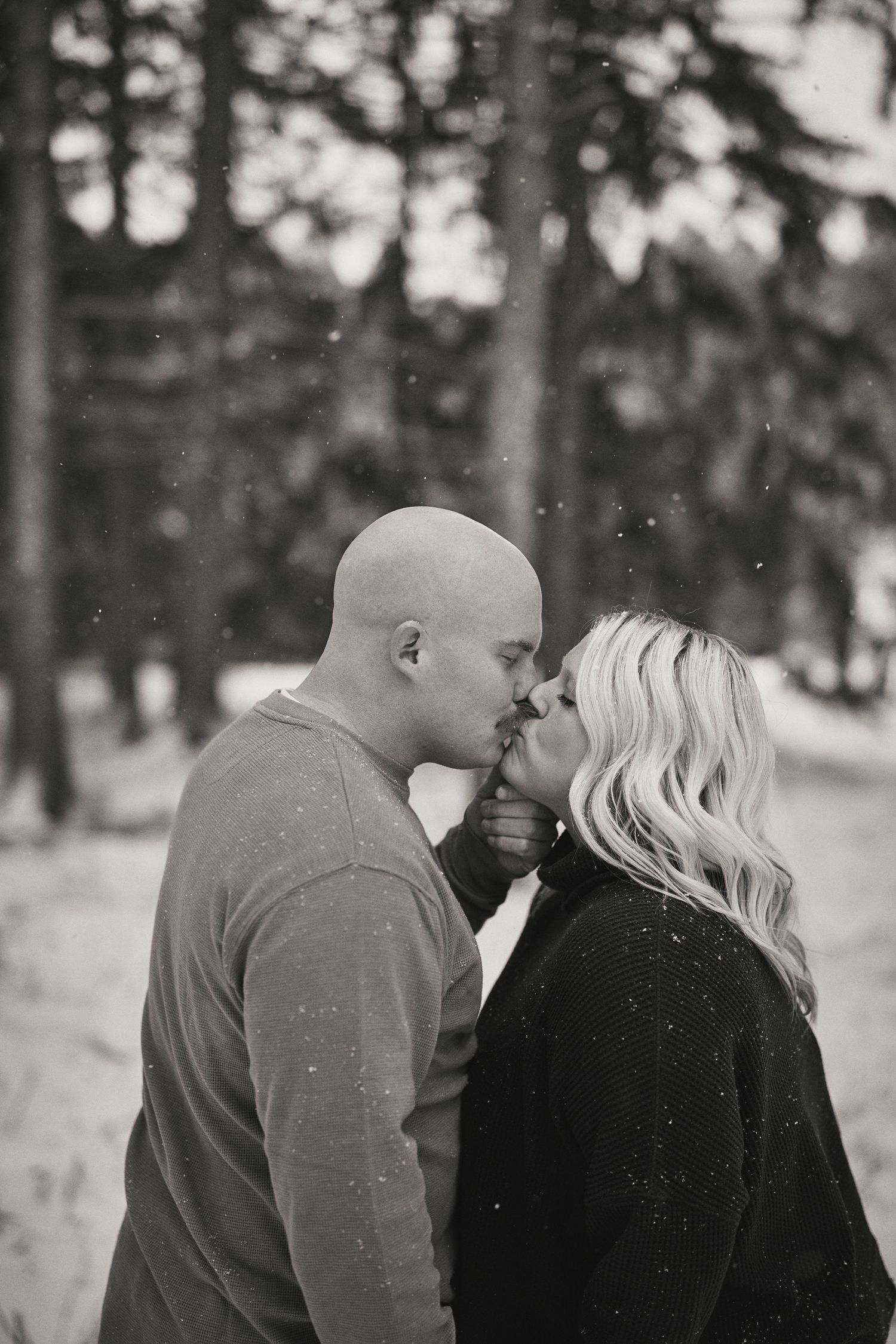 A romantic black and white portrait of a couple in winter attire sharing an intimate moment among snowy trees.