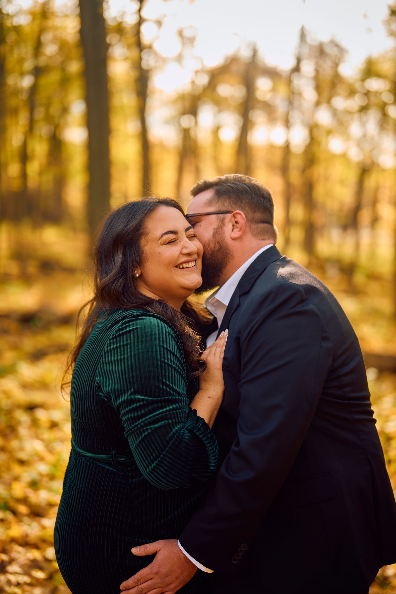 A romantic autumn engagement photo shows a couple embracing among fallen leaves in a golden-lit forest setting.
