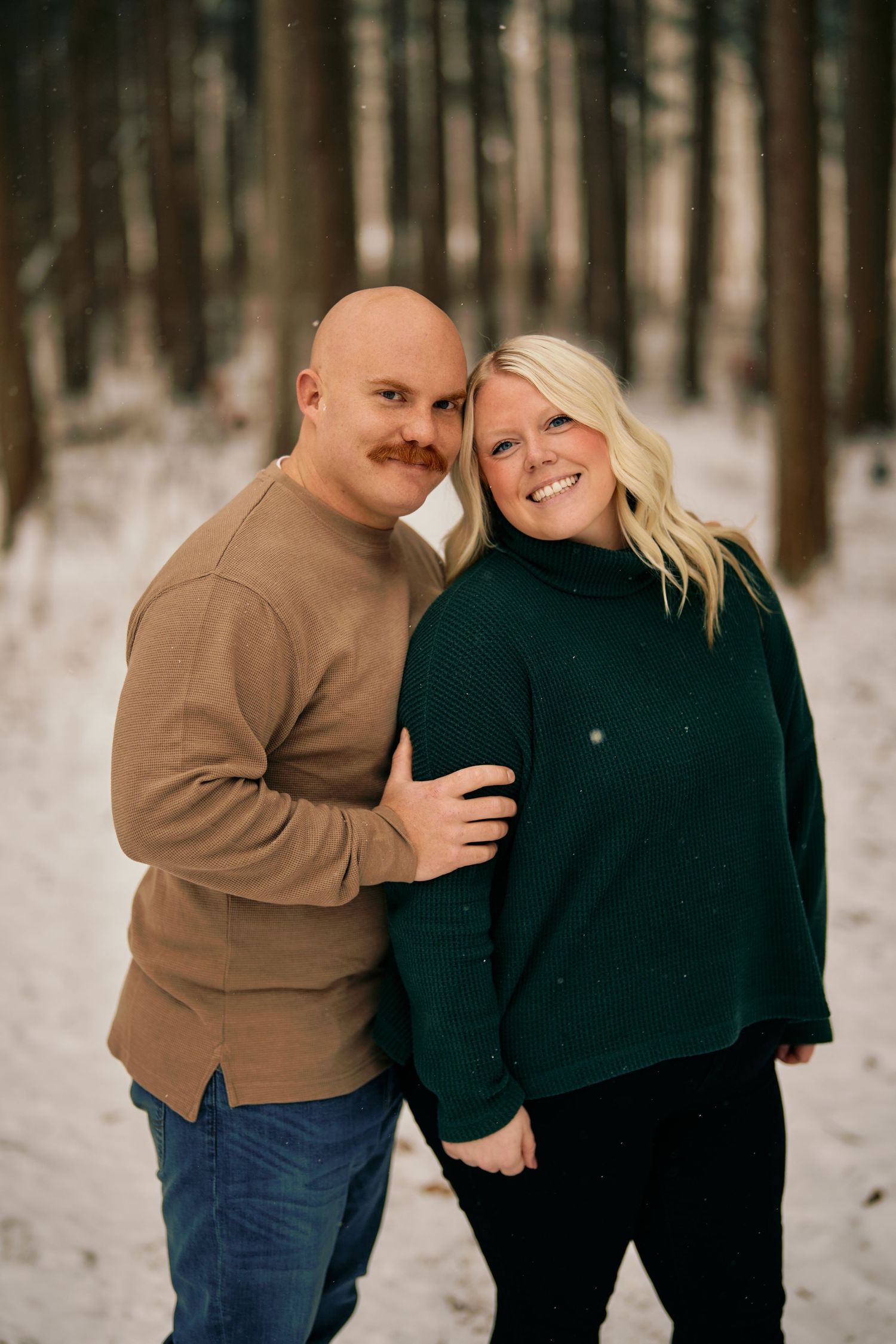 A couple poses together in a snowy forest setting wearing cozy winter sweaters in brown and dark green.