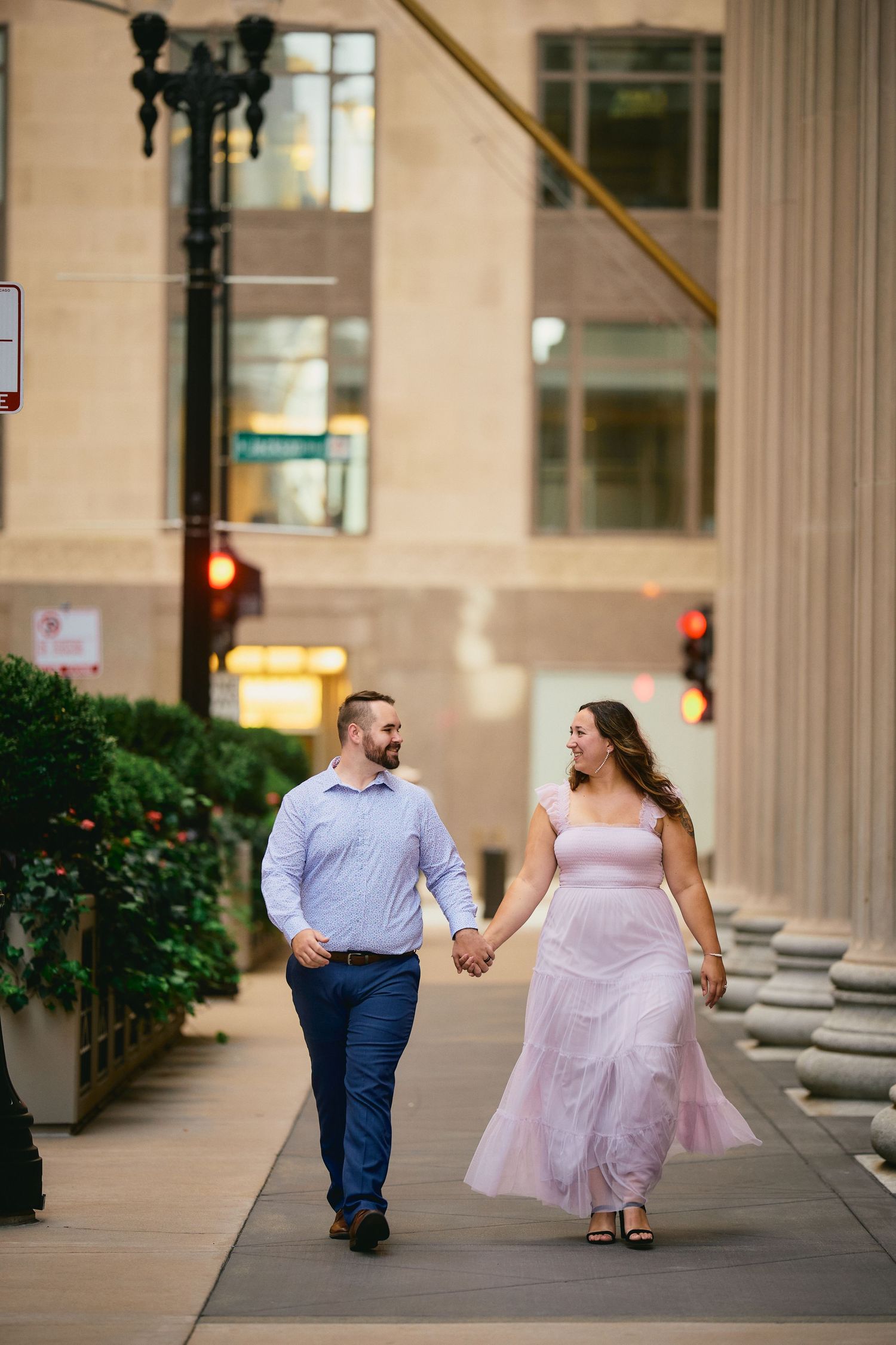 A couple walks hand in hand along a city sidewalk at dusk, with her wearing a flowing pink dress and him in business casual.