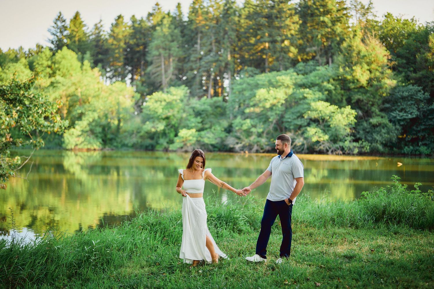 Romantic couple holding hands by a serene lake surrounded by lush green forest on a summer evening.