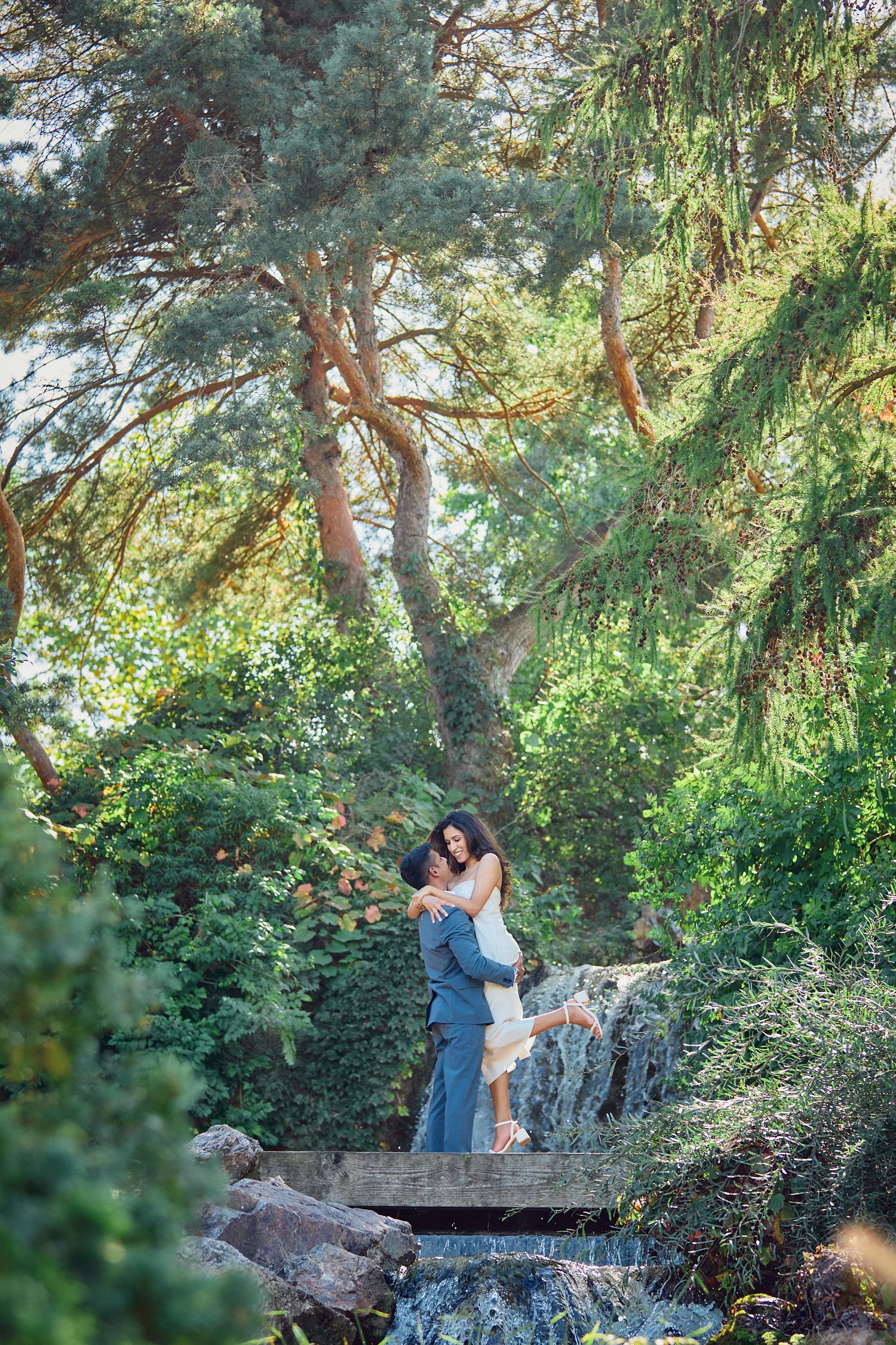 A couple shares a romantic moment on a wooden bridge surrounded by lush greenery and a waterfall.