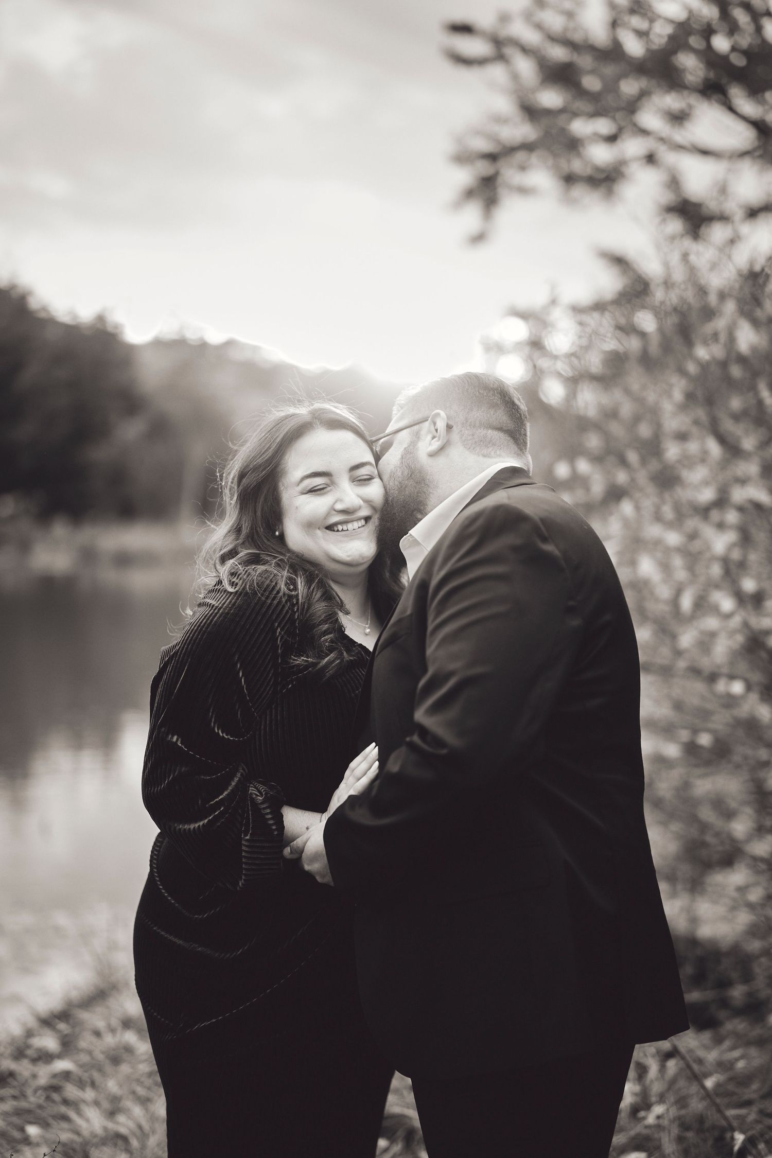 A romantic couple in formal attire share an intimate moment by a lake during a black and white photoshoot.