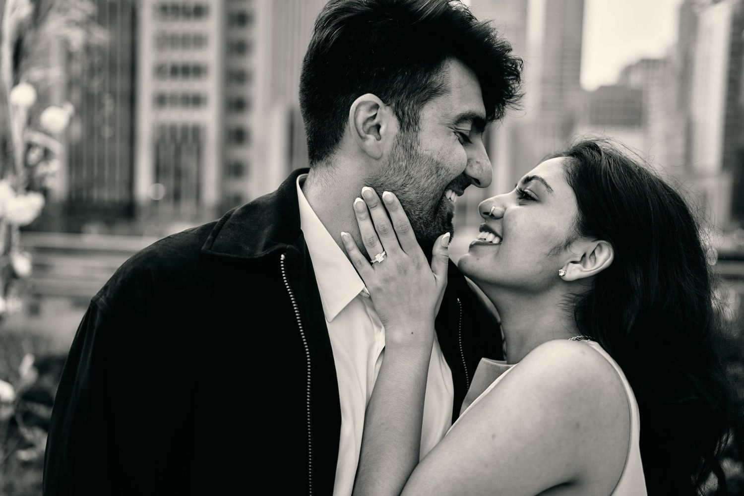 A romantic black and white portrait of a couple sharing an intimate moment in an urban setting with city buildings in background.