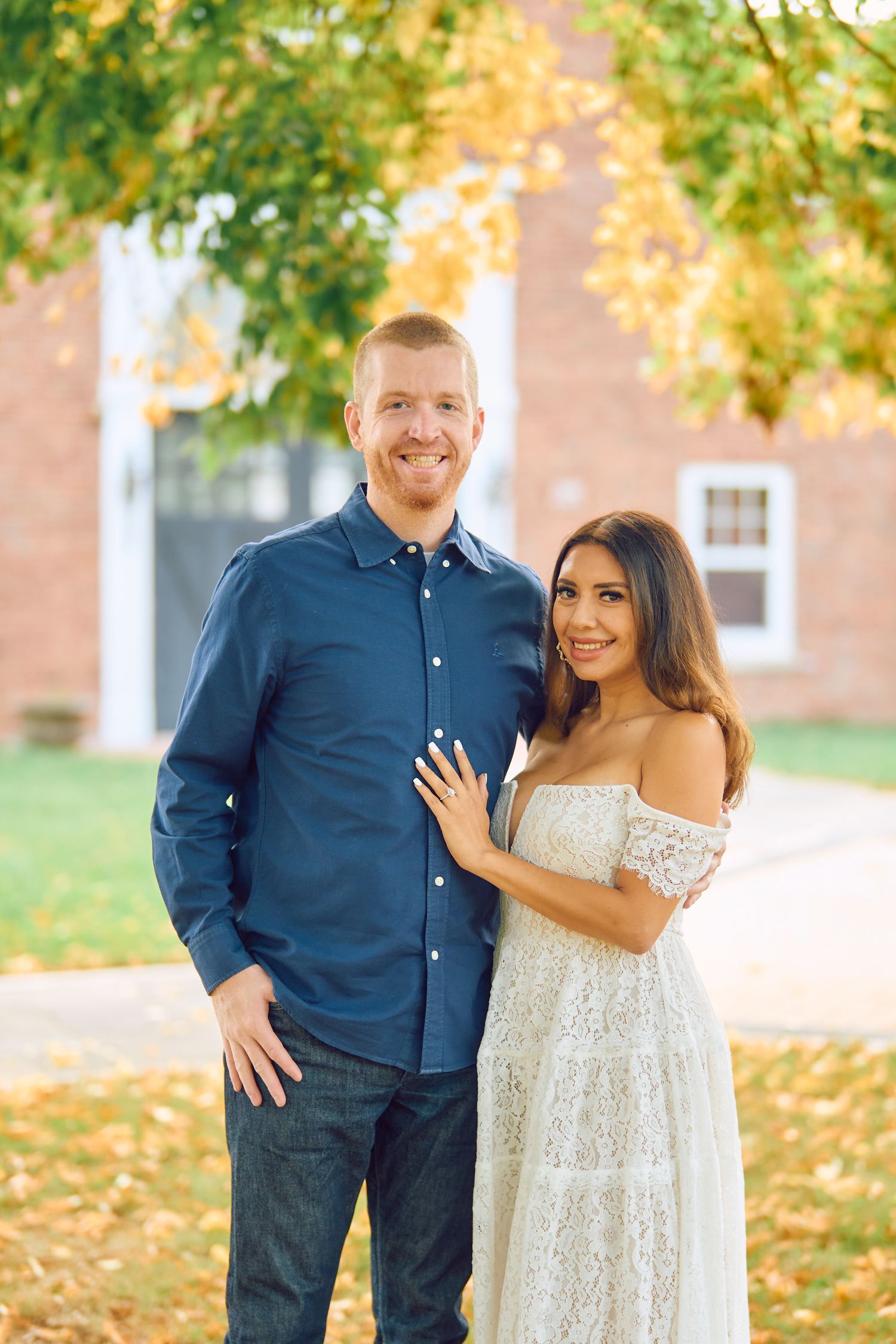 A couple poses for autumn engagement photos outdoors with fall foliage and a brick building in the background.