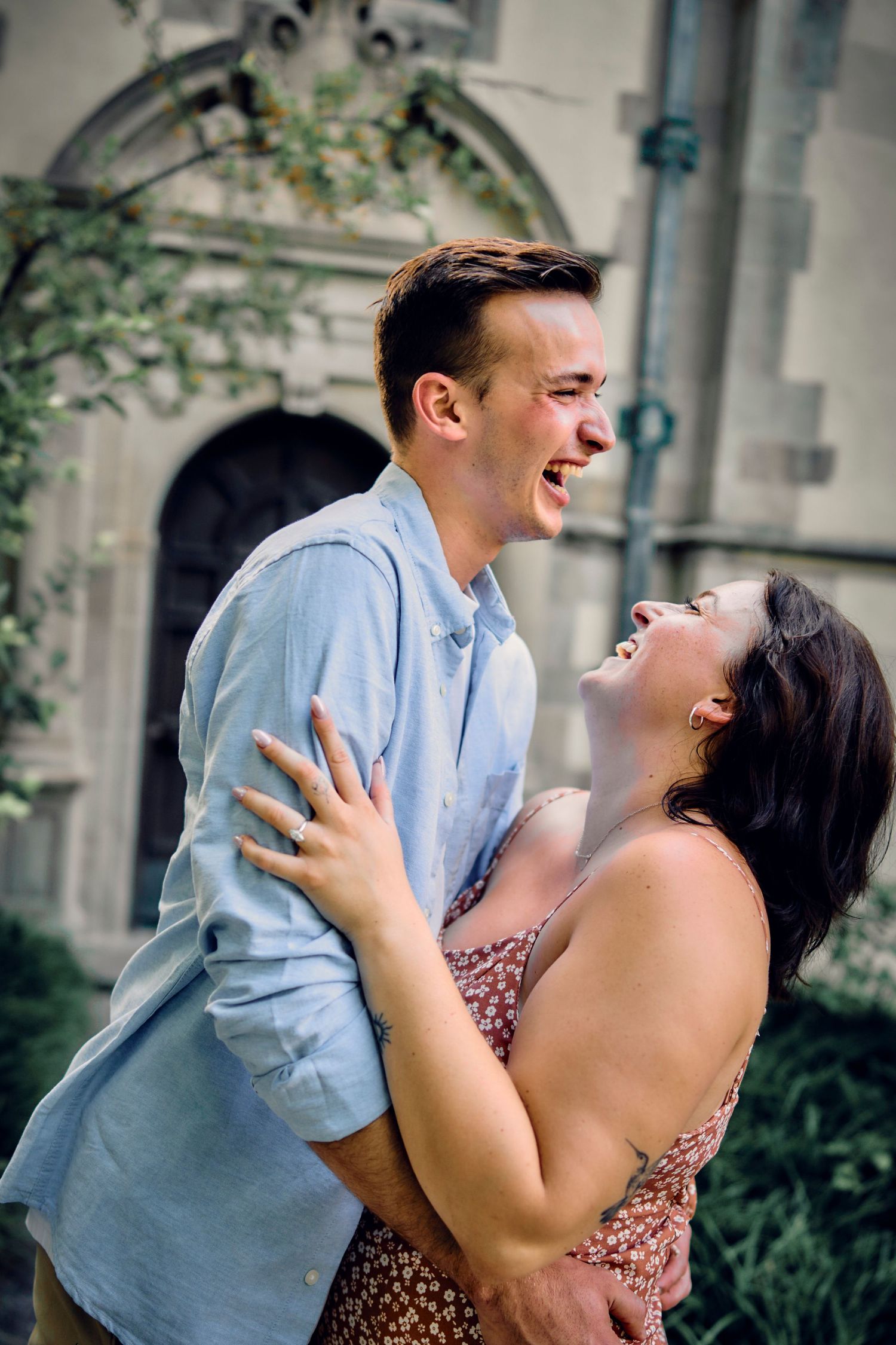 A couple shares a joyful moment of laughter together against a stone building backdrop with greenery.