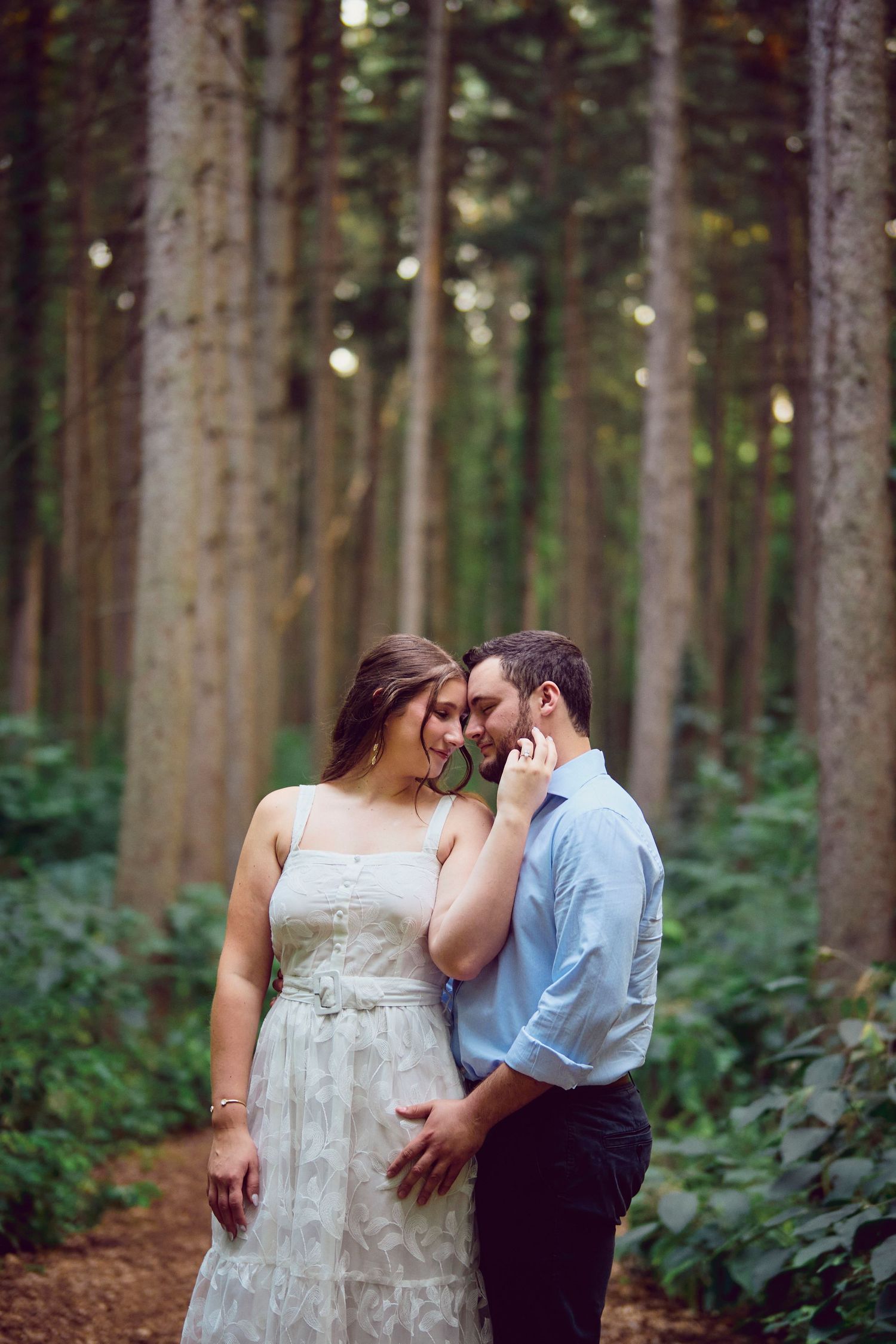 A tender romantic moment between a couple in formal attire standing in a serene forest setting.