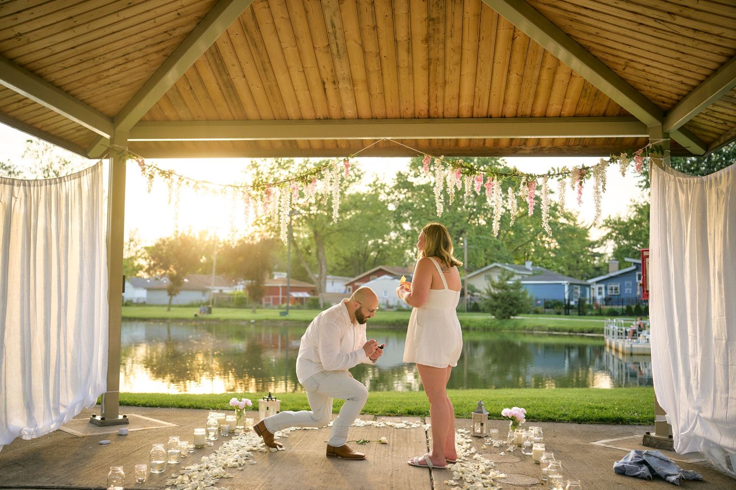 A romantic proposal scene at sunset under a wooden gazebo by a serene lake with white curtains billowing in the breeze.