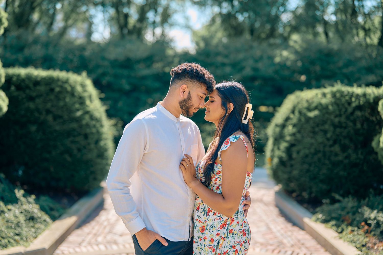 A couple shares an intimate moment in a garden pathway lined with manicured hedges on a sunny afternoon.
