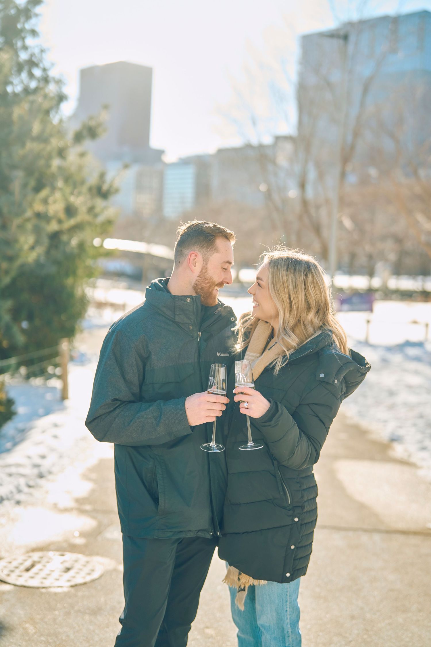A couple shares champagne outdoors on a winter day with snowy city buildings in the background.