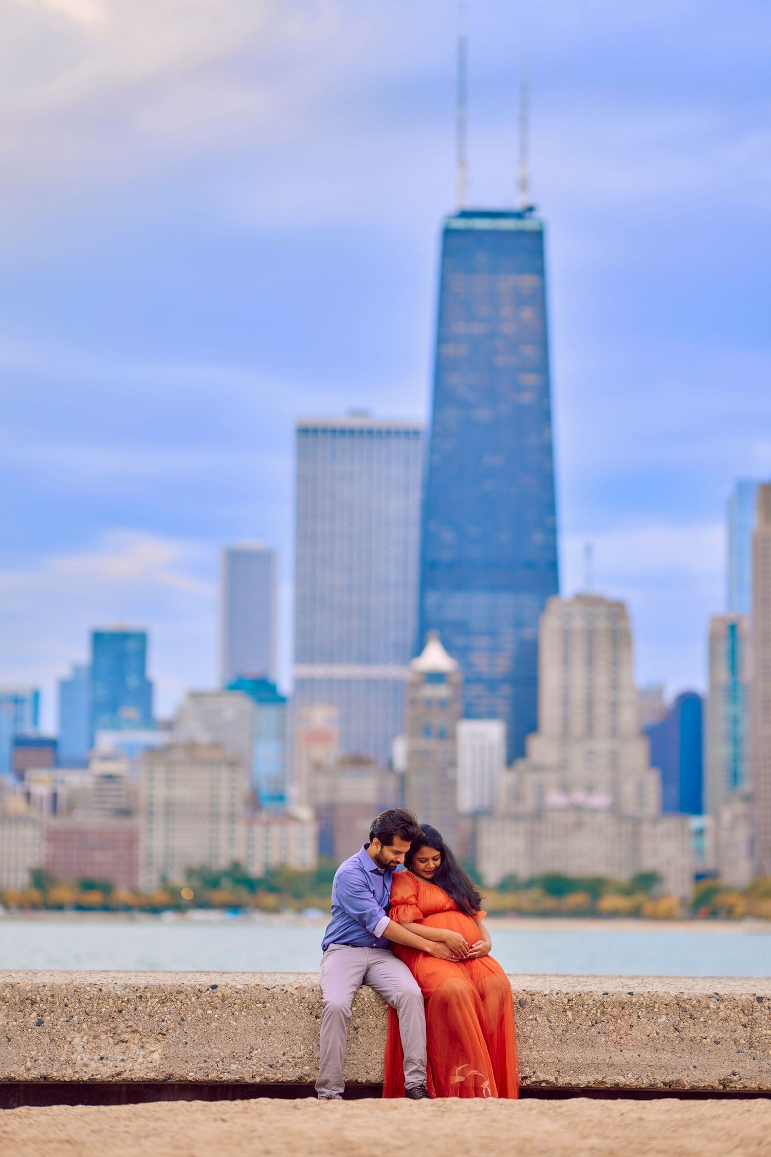 A couple sits together on a waterfront wall with Chicago's iconic John Hancock Center and skyline in the background.