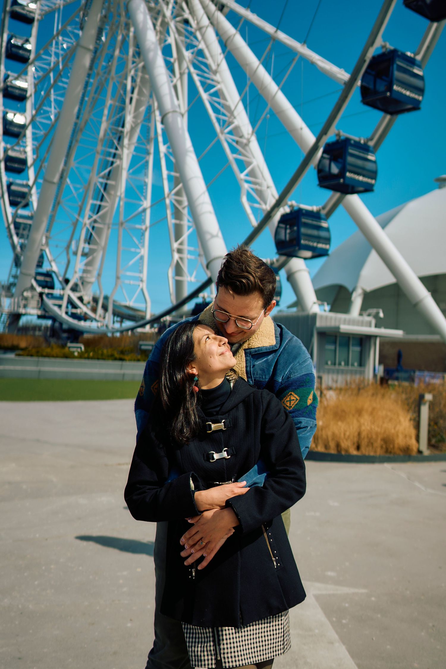 A romantic couple embraces in front of a large ferris wheel on a sunny day with bright blue skies.