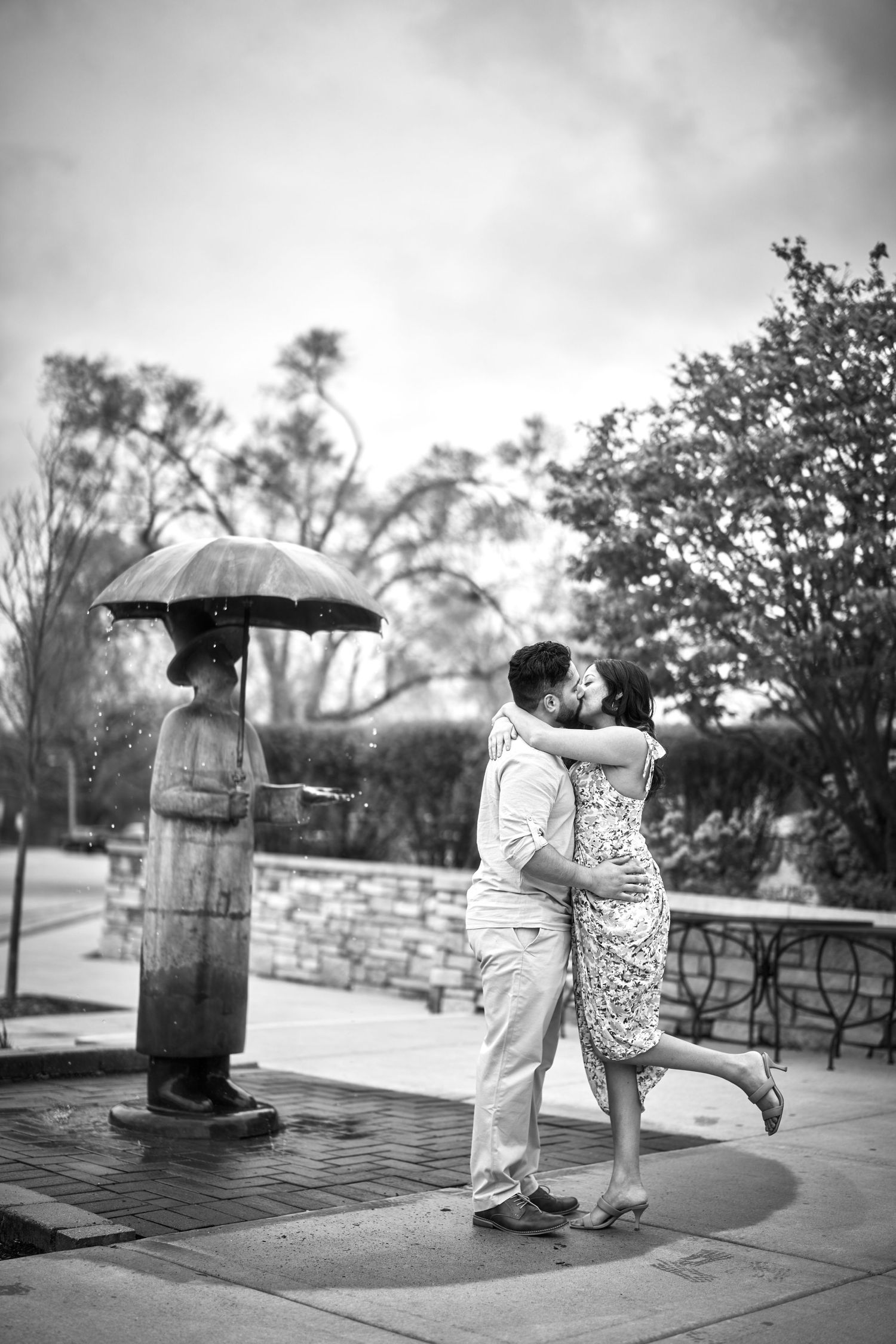 A romantic black and white photo of a couple embracing on a rainy day beside a decorative metal umbrella statue.
