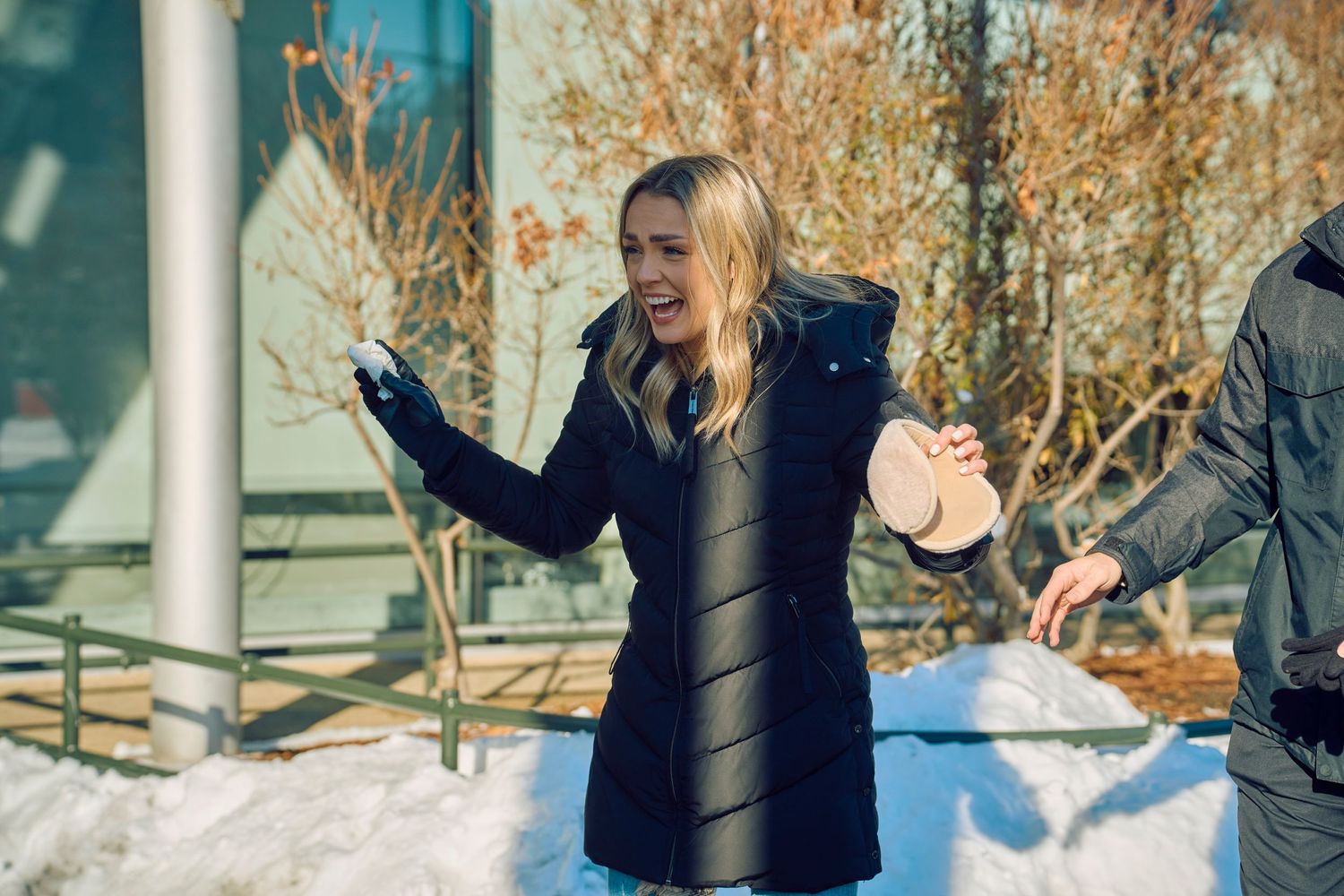 Someone in a puffy black winter coat gestures excitedly while standing in a snowy outdoor setting.