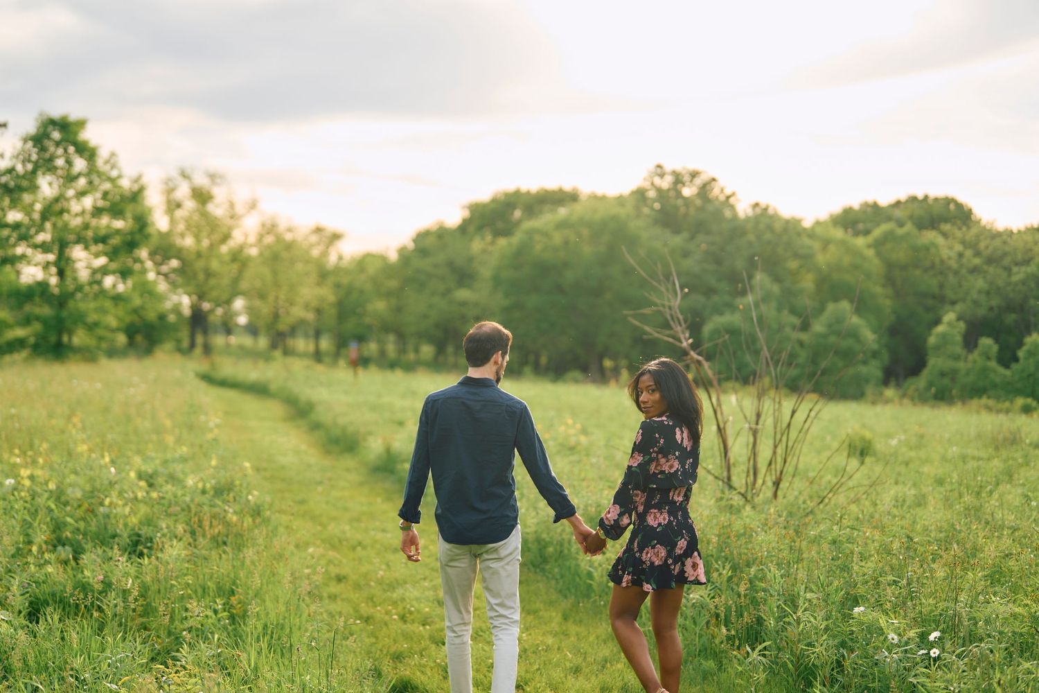 Couple walking hand in hand through a sunlit meadow with wildflowers and trees in the background.