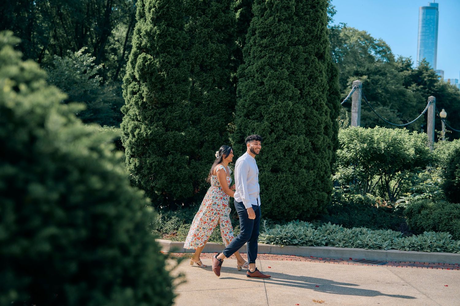 Two people stroll along a garden path lined with tall cypress trees and greenery on a sunny day.