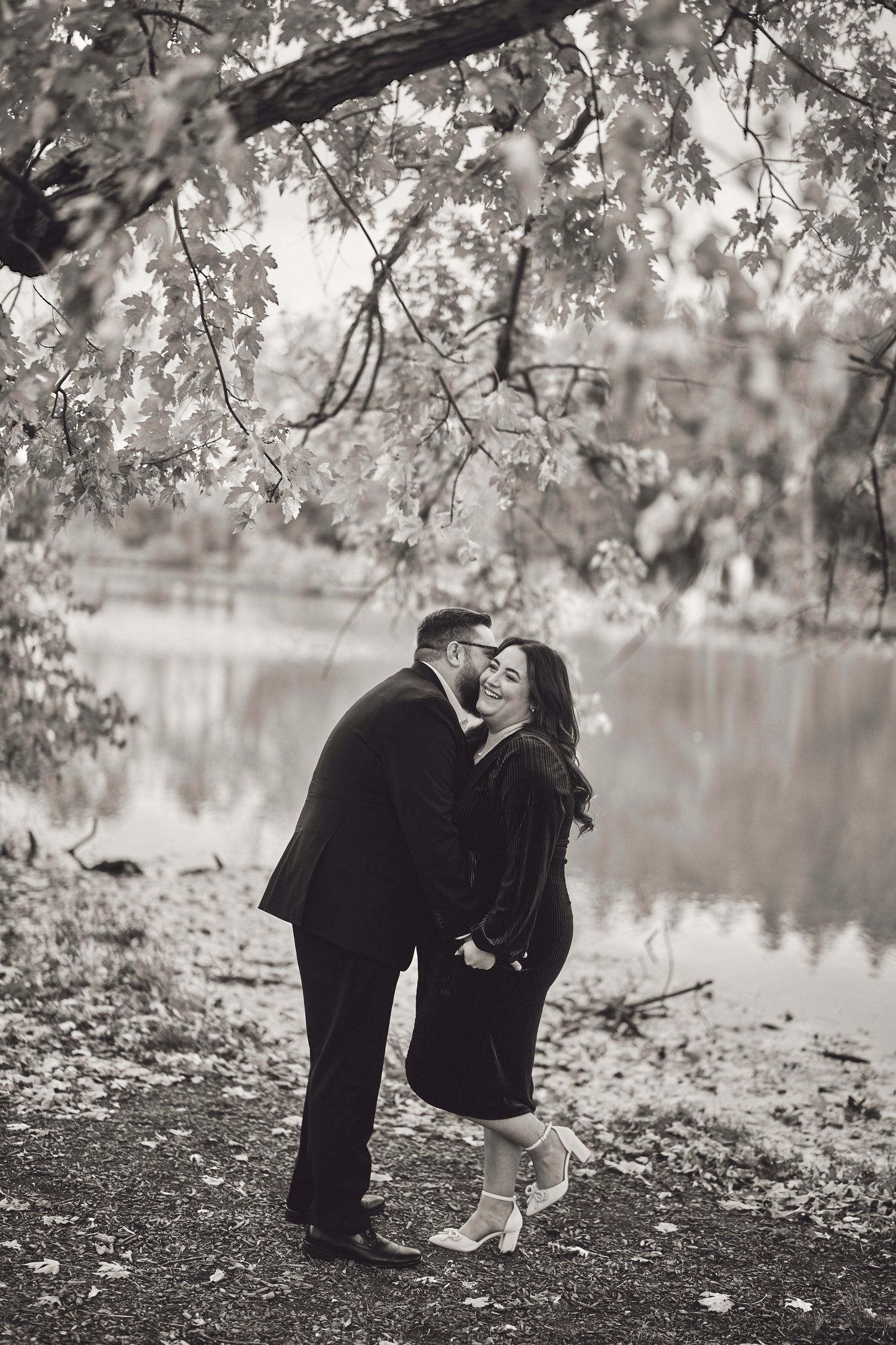 A romantic couple shares a tender moment under blossoming trees by a tranquil lake in black and white photography.