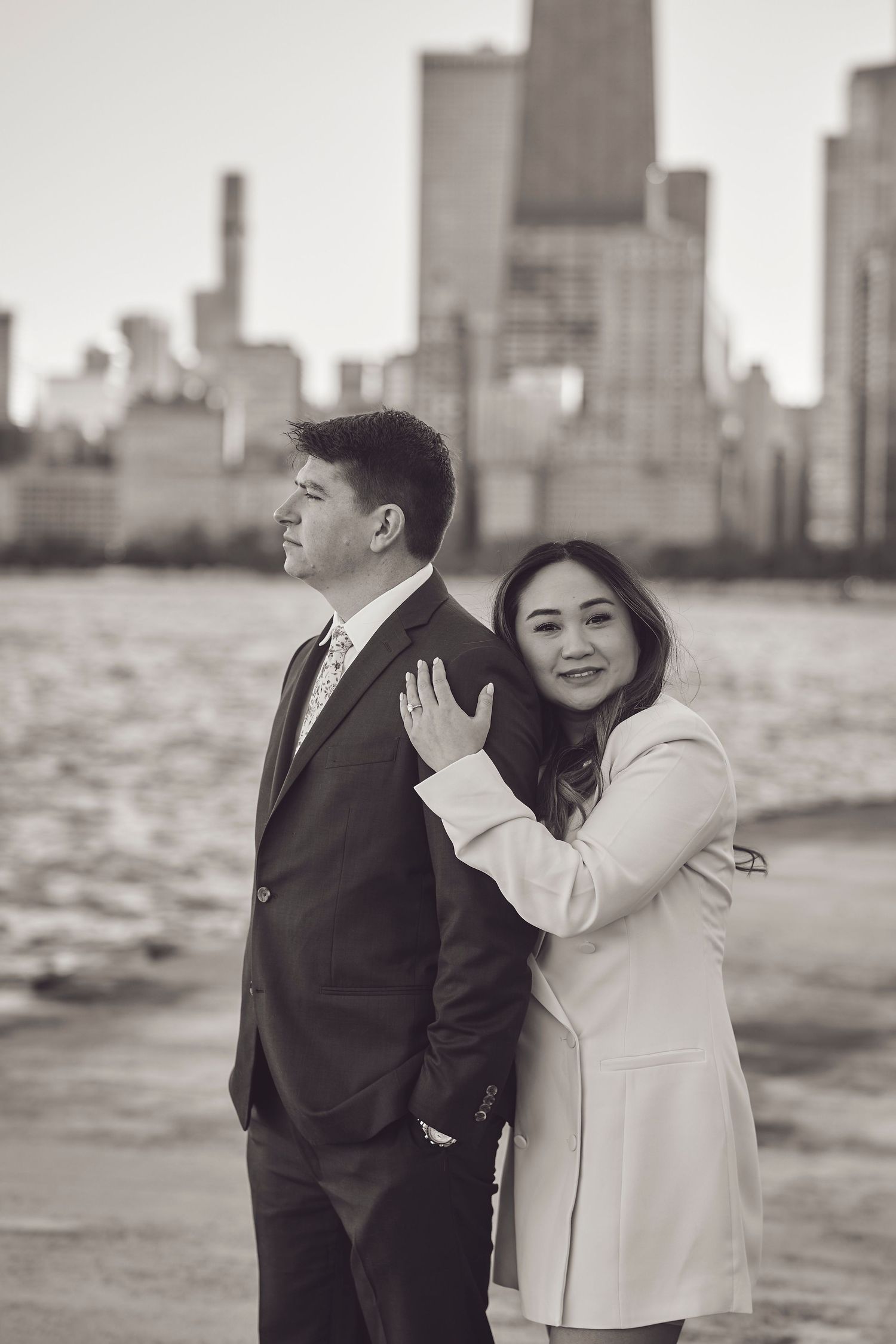 A couple embraces on the waterfront with a cityscape silhouette in the background during a black and white photoshoot.