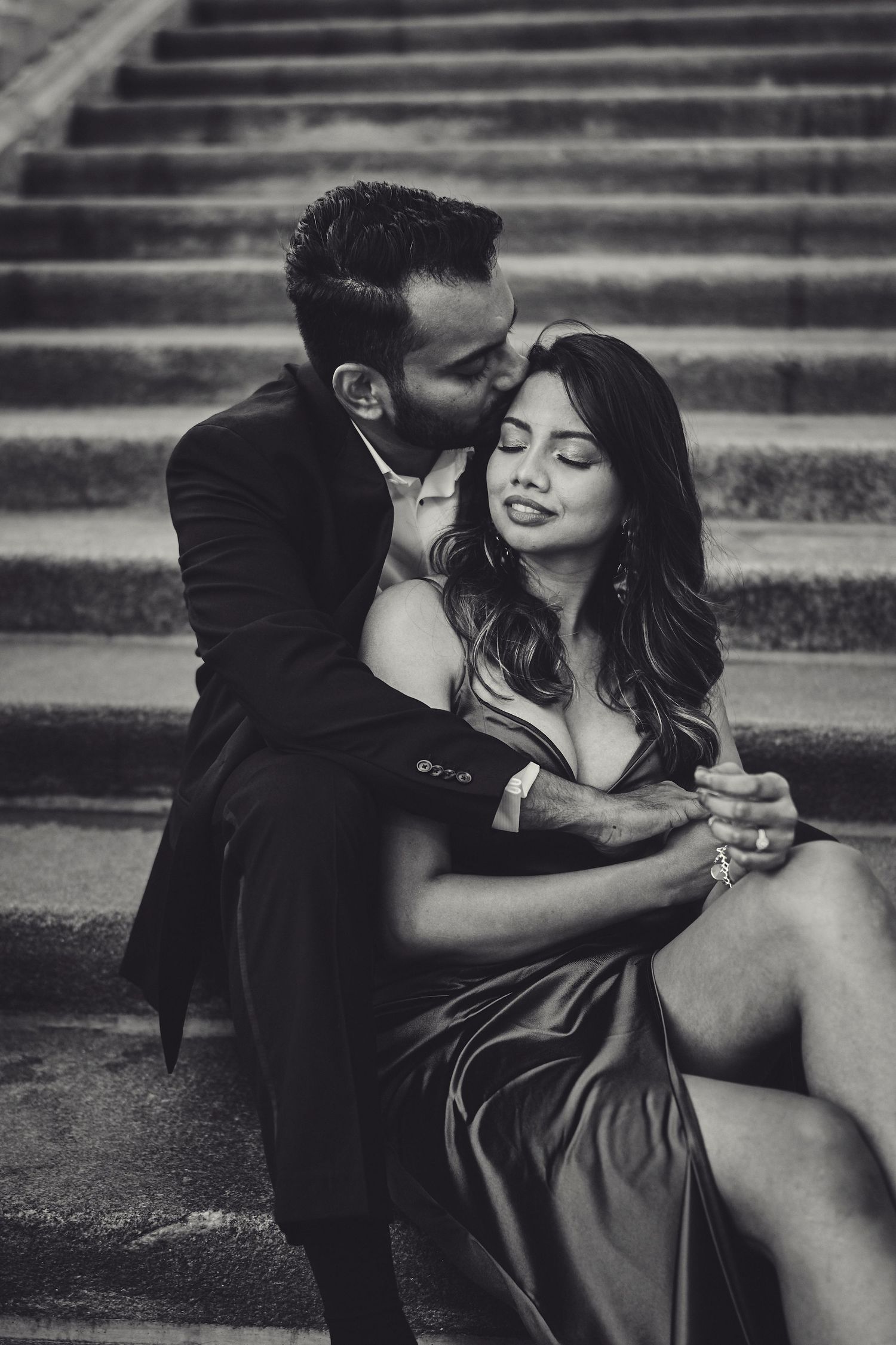 Romantic black and white portrait of a couple embracing on stone steps, with soft natural lighting and elegant attire.
