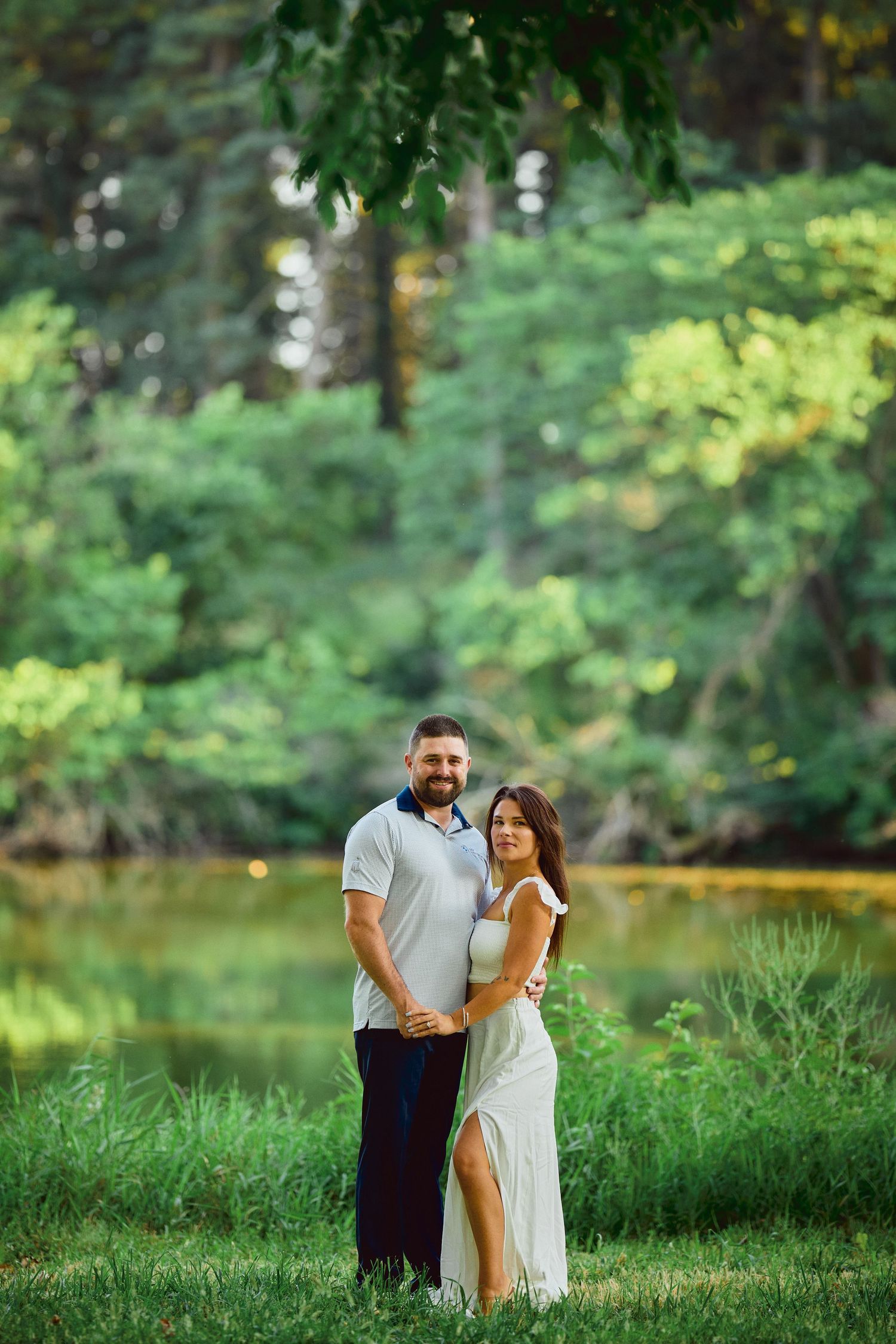 A happy couple poses together outdoors in a romantic natural setting with lush greenery and a lake in the background.