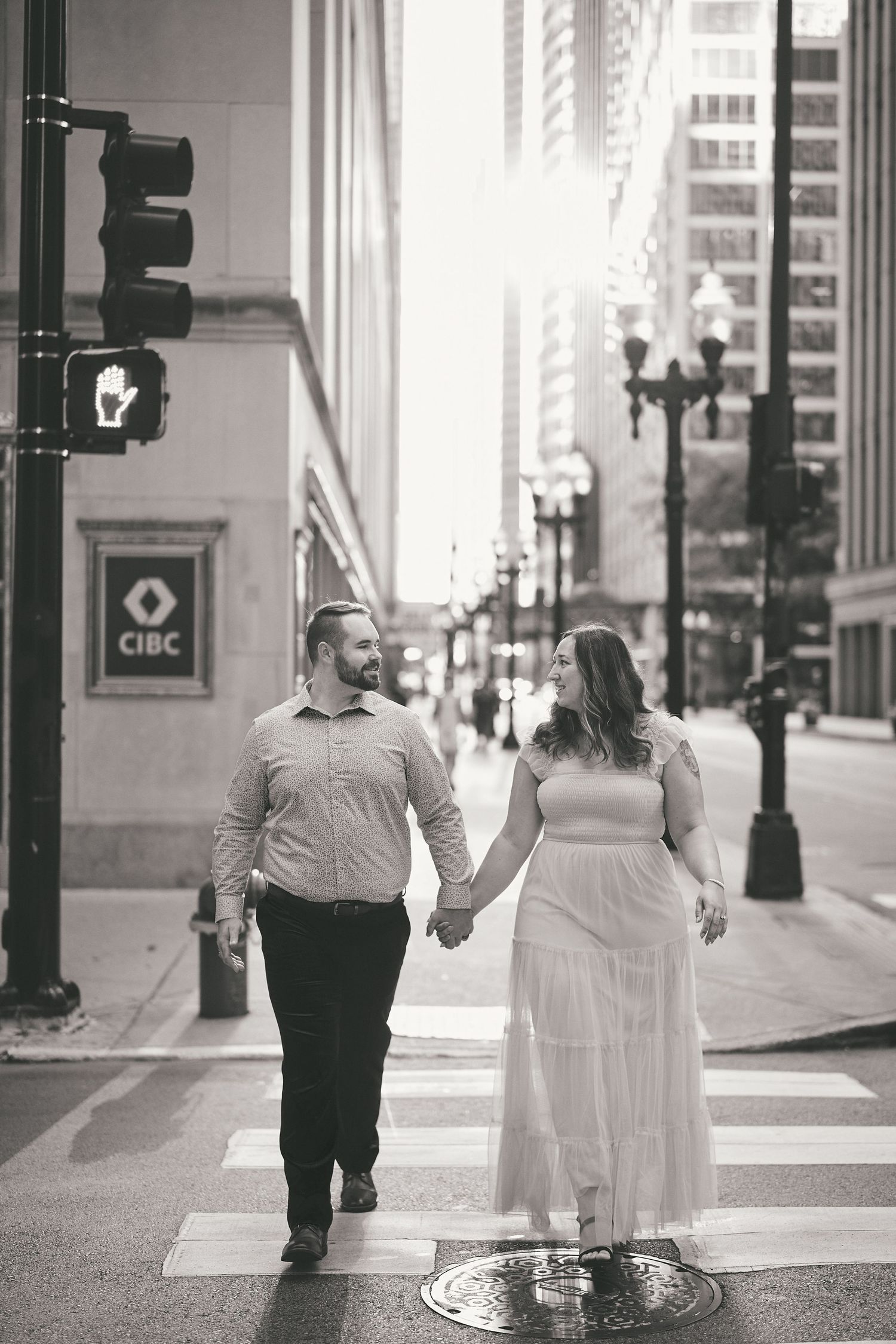 A couple holds hands while crossing a downtown street with a CIBC bank sign and city lampposts in black and white.