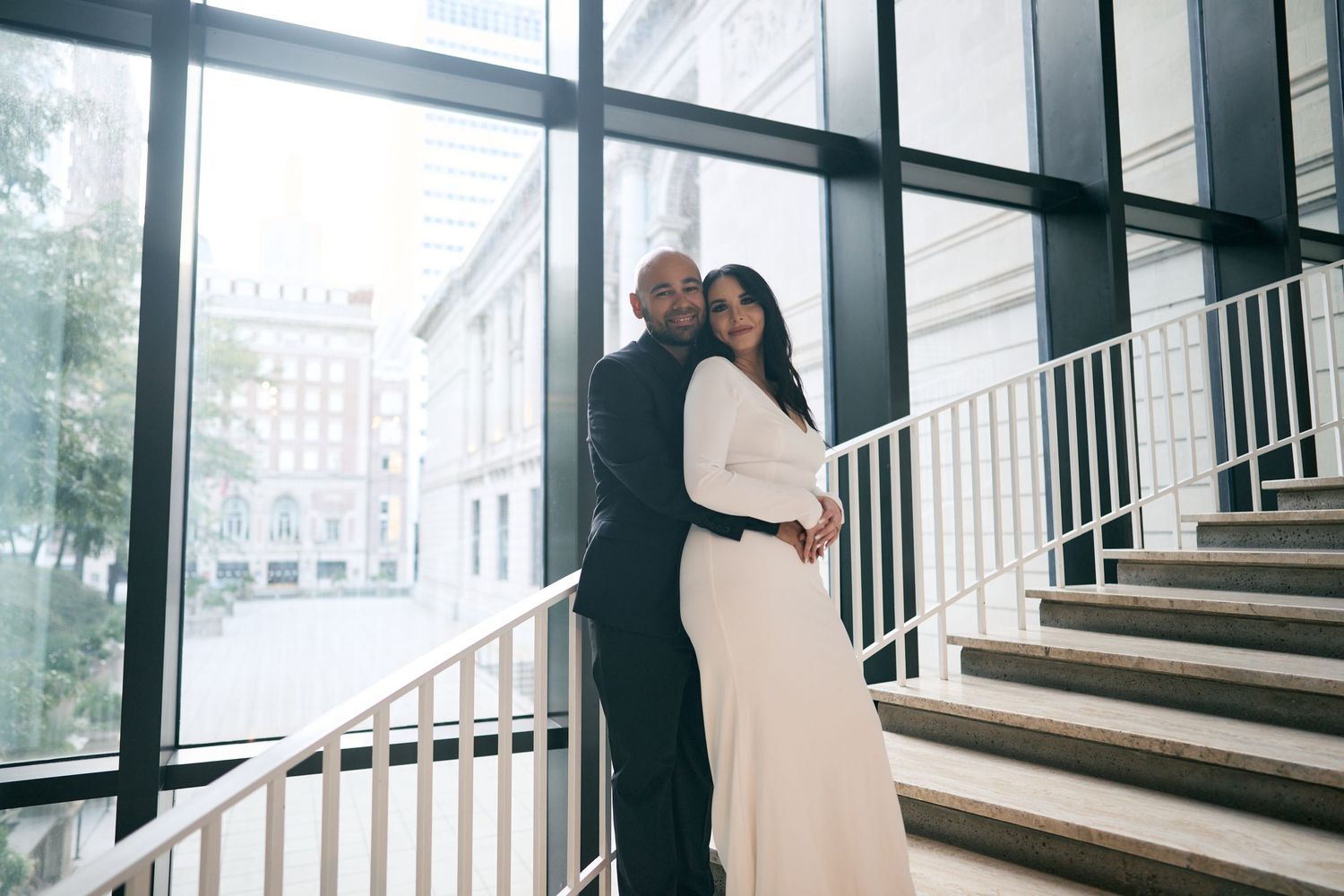 A couple embraces on modern glass staircase with an urban backdrop visible through large windows.
