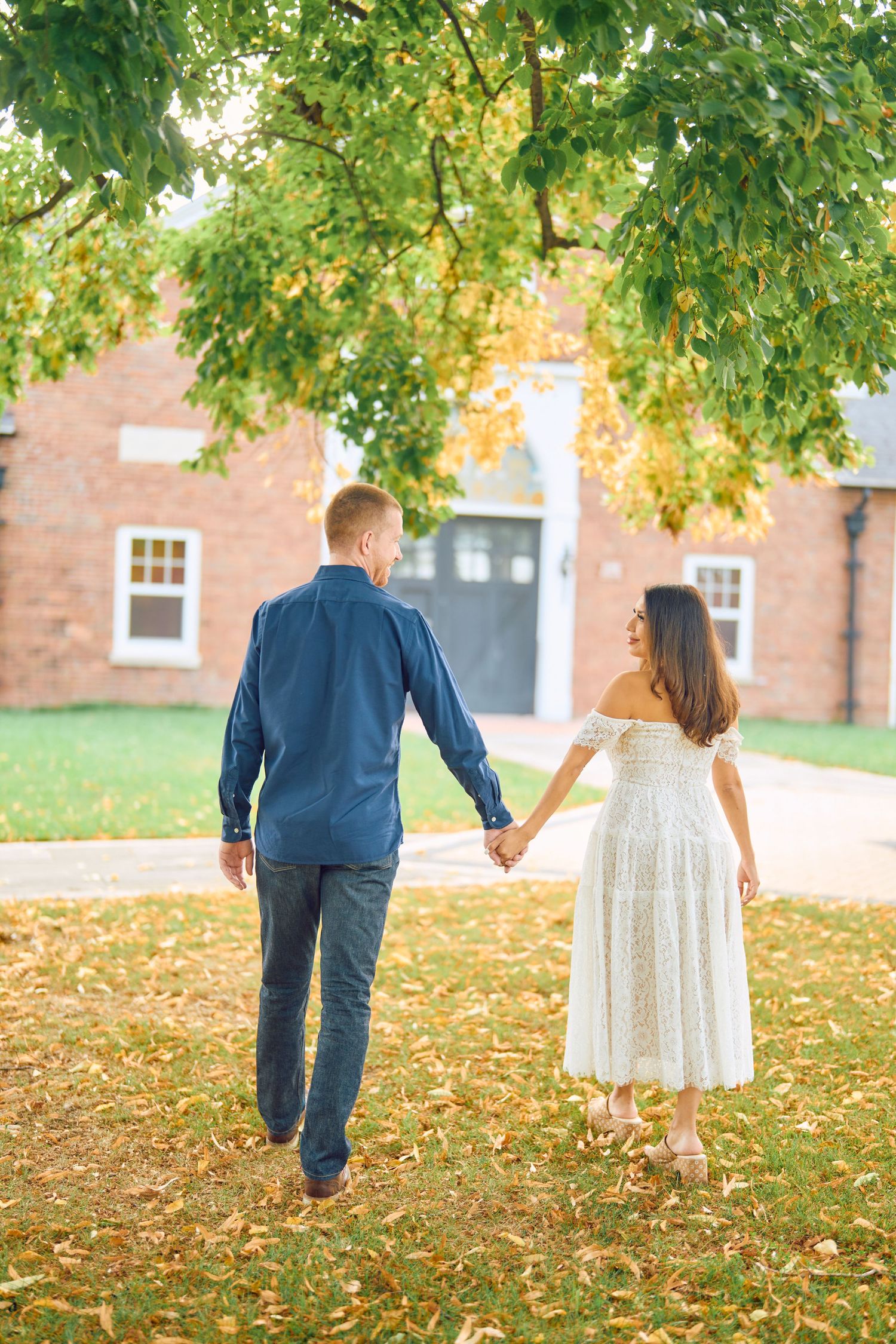 A couple walks hand in hand on fallen autumn leaves near a brick building in warm sunlight.