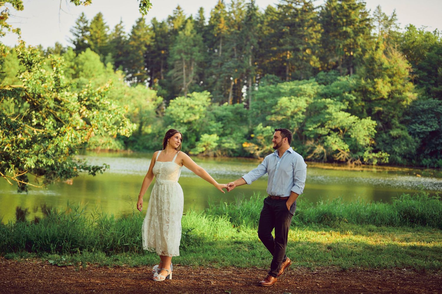 A couple in formal attire walks hand in hand along a lakeside path surrounded by lush summer greenery.