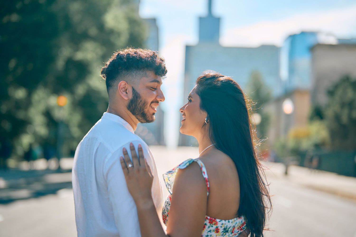 A couple shares a romantic moment on a sun-lit urban street with blurred city buildings in the background.
