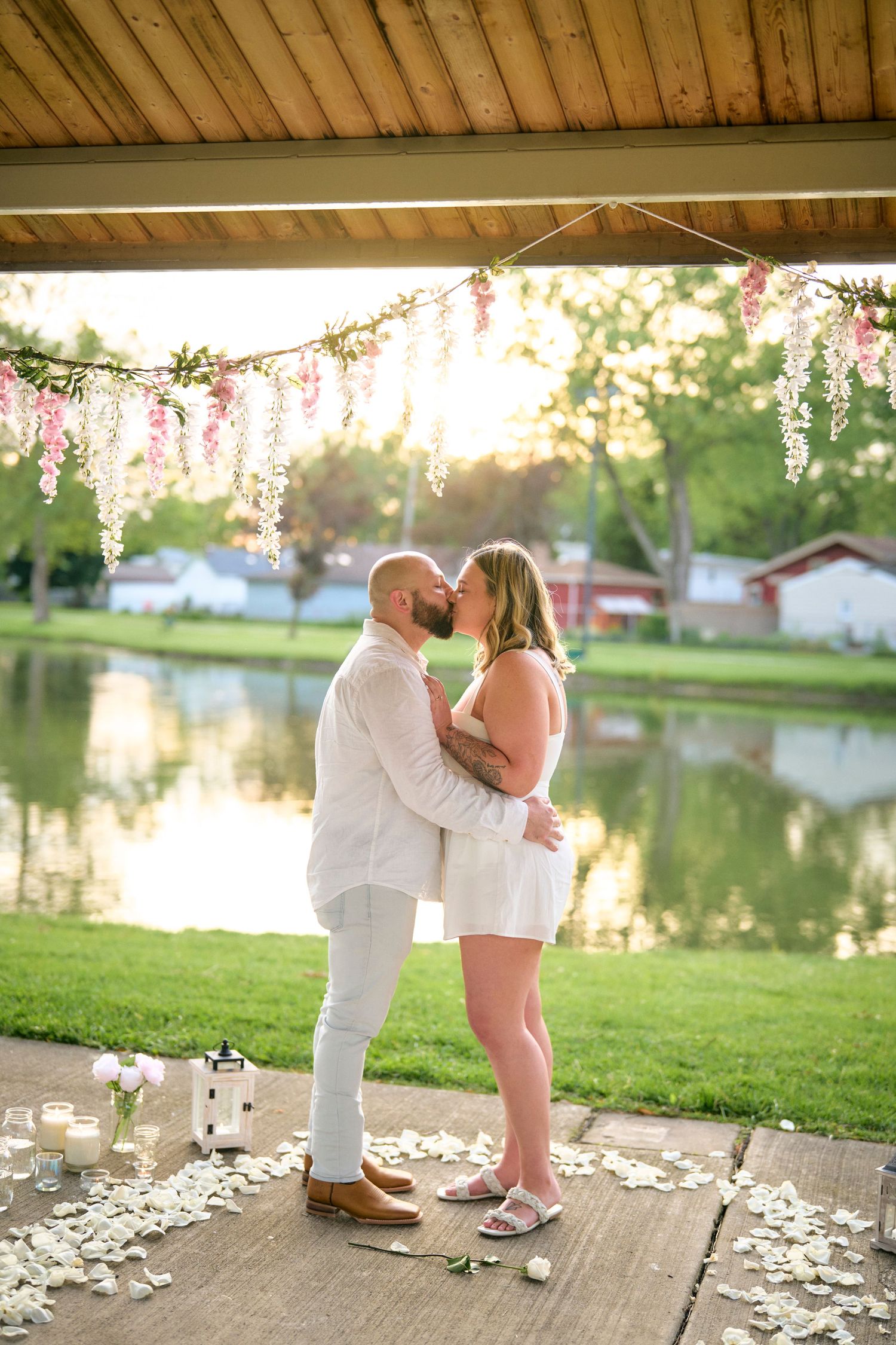A romantic couple shares an intimate moment under floral decor by a lake at sunset, petals scattered on the ground.