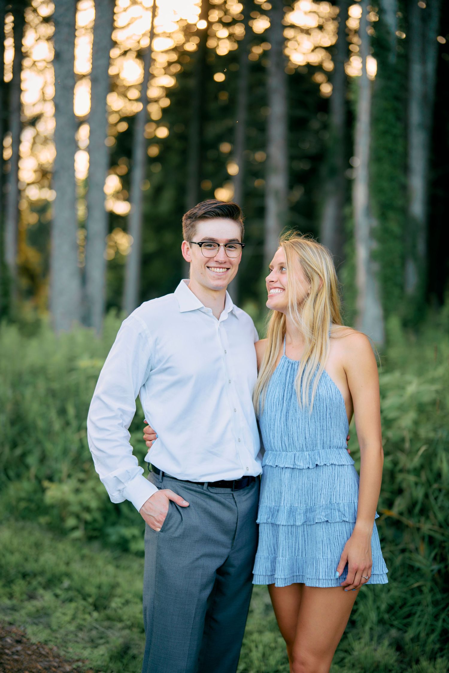 A couple in formal attire shares a happy moment together in a sun-dappled forest during golden hour.