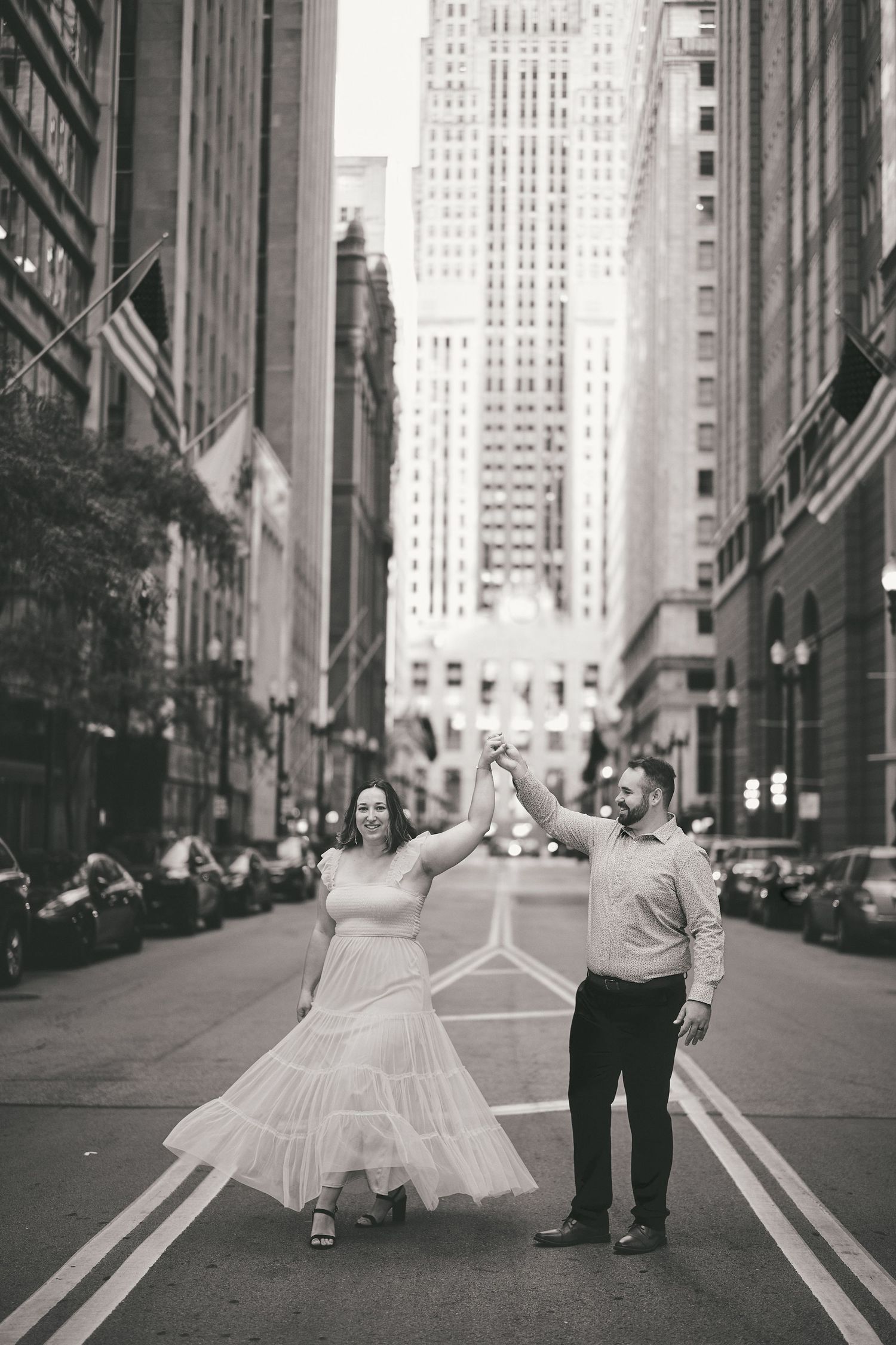 A couple dances joyfully in the middle of a city street with tall buildings and American flags in the background.