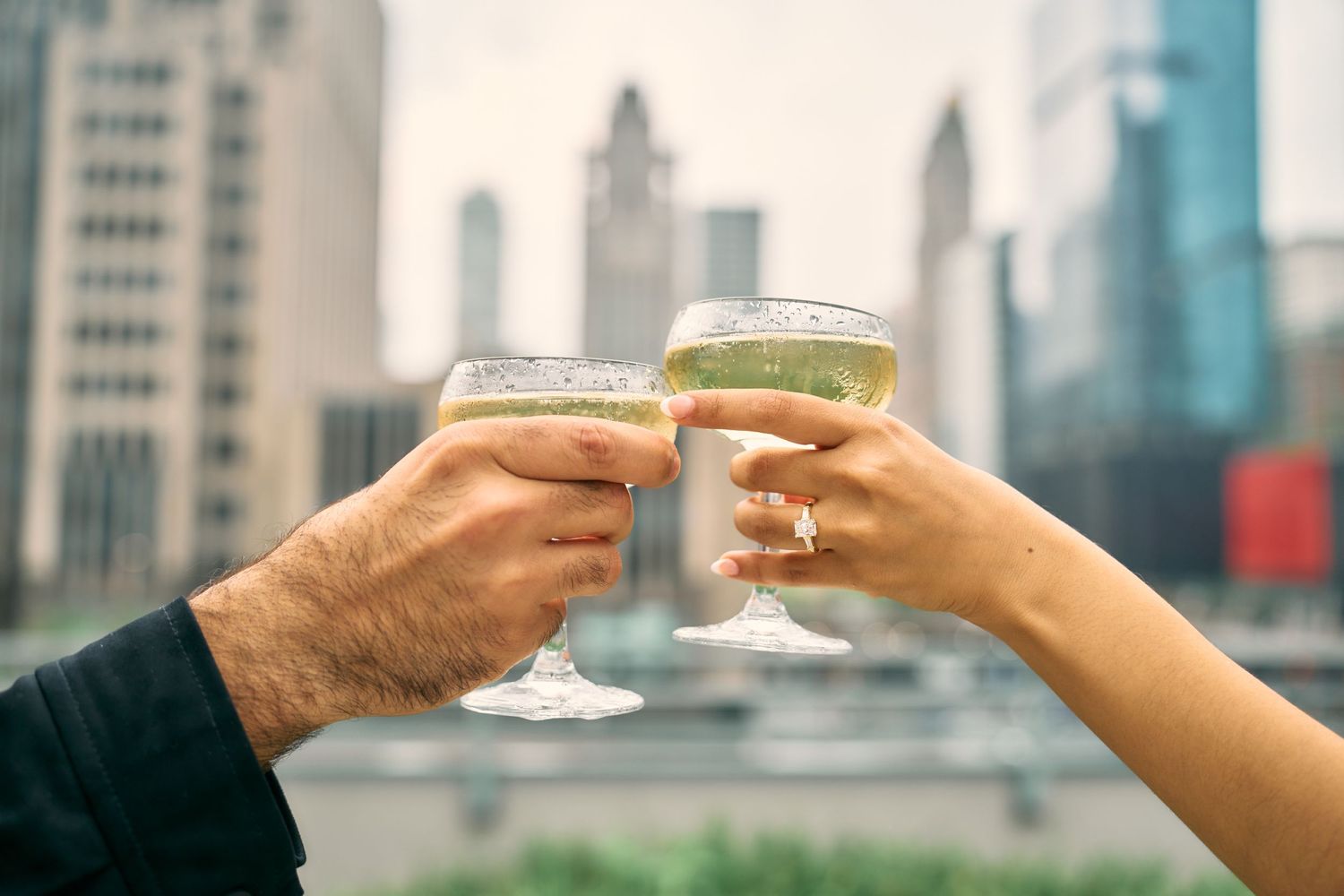 Two hands toasting with wine glasses against a blurry city skyline background in golden afternoon light.