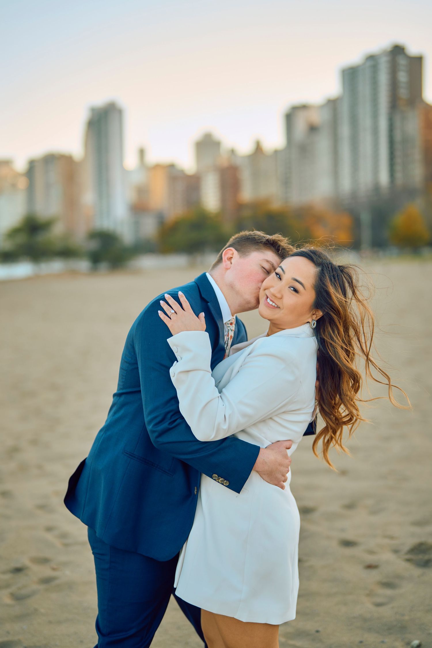 Happy couple embracing on sandy beach with city skyline during sunset, showing joyful expressions and casual outfits.