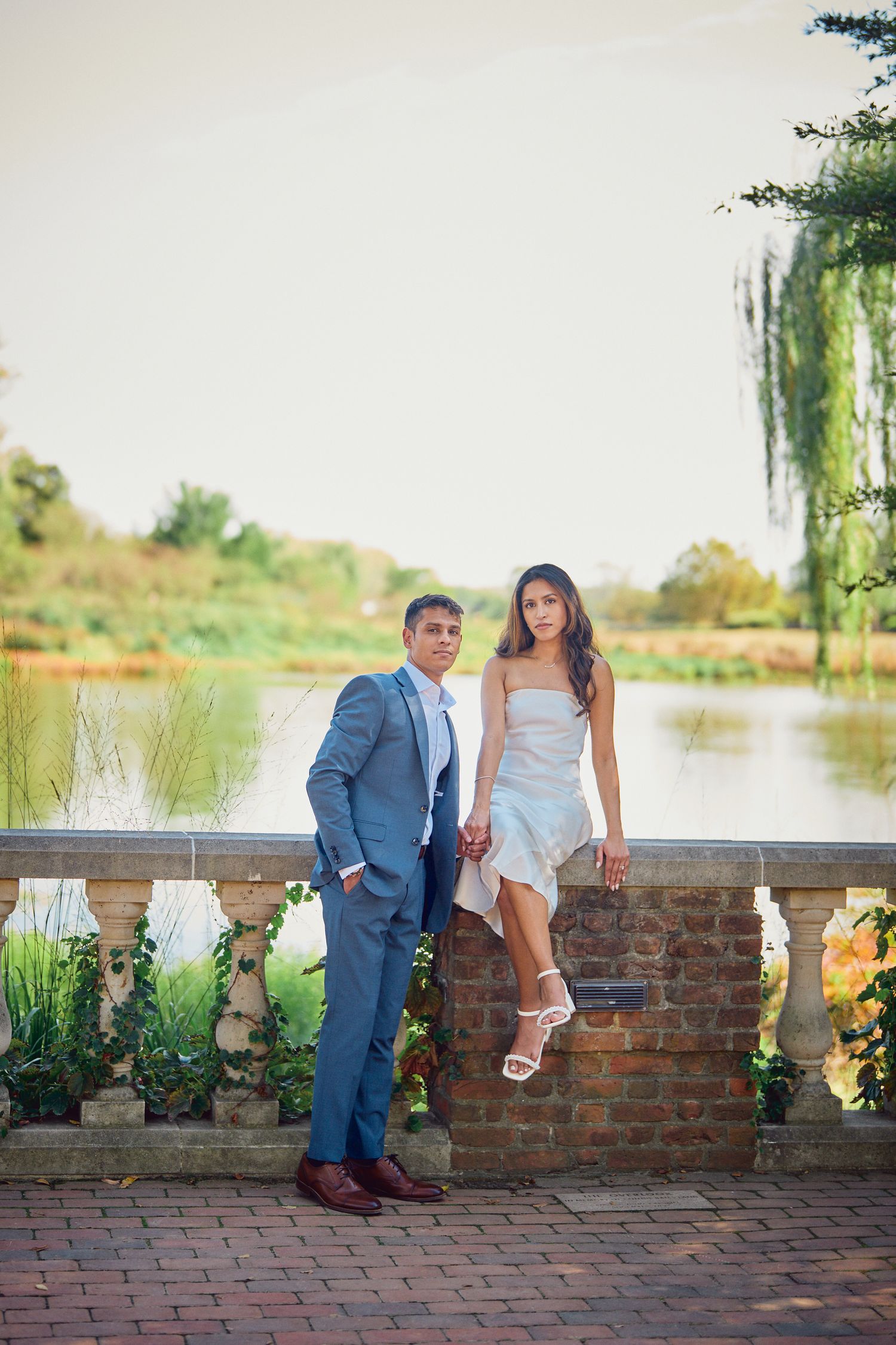 Couple poses elegantly by a lake on brick terrace with flowering plants and willow tree in background.
