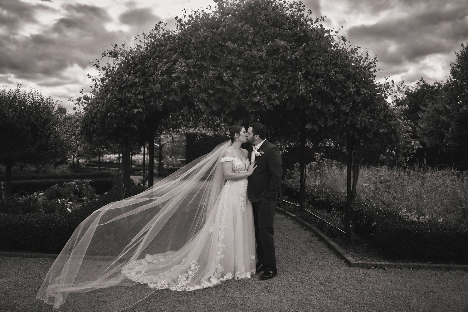 Dramatic black and white wedding photo with flowing veil captured in a garden path beneath arching trees.