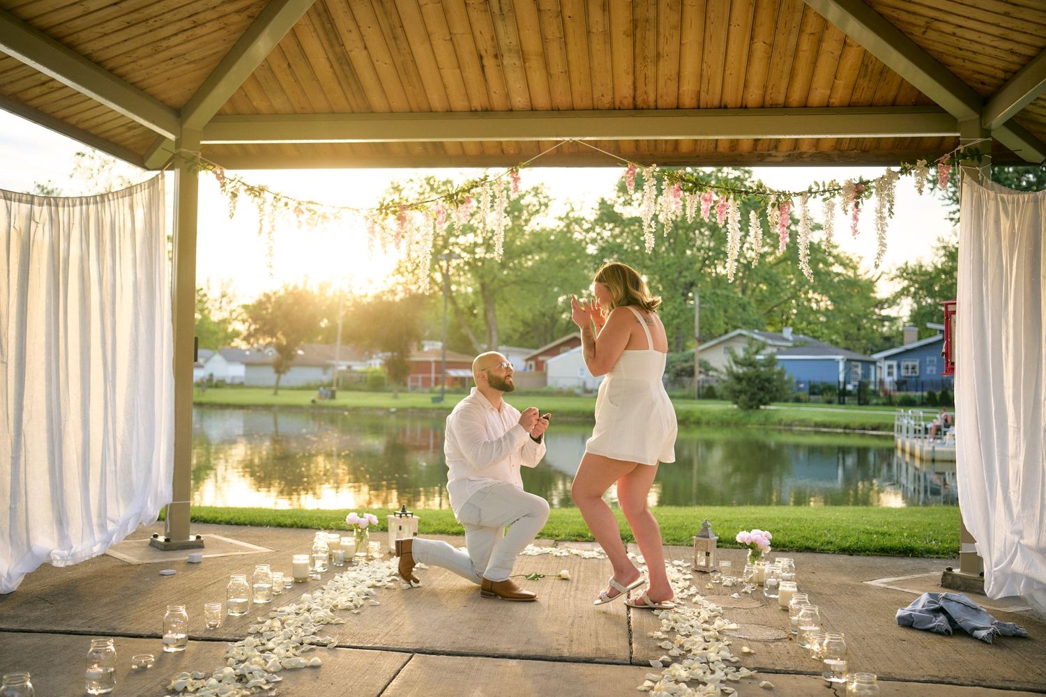 A romantic sunset proposal moment in a gazebo decorated with candles beside a peaceful lake.