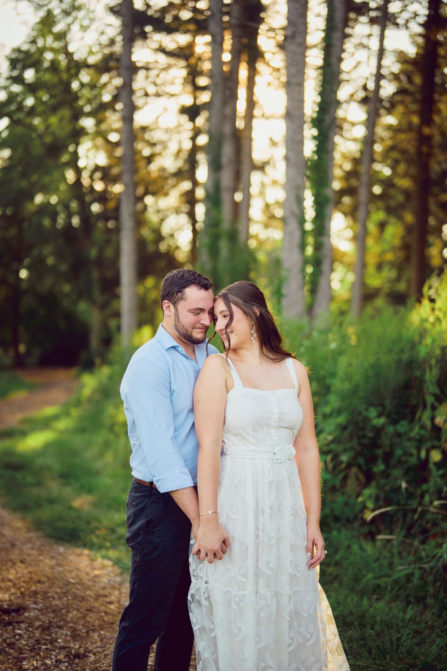 Romantic couple in formal attire share an intimate moment on a sunlit forest path during golden hour.