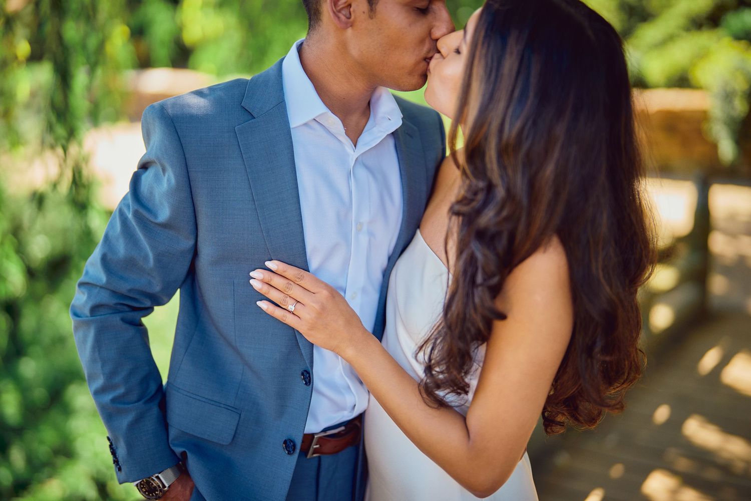 A couple dressed in blue suit and white dress embrace intimately in a sunny garden setting.