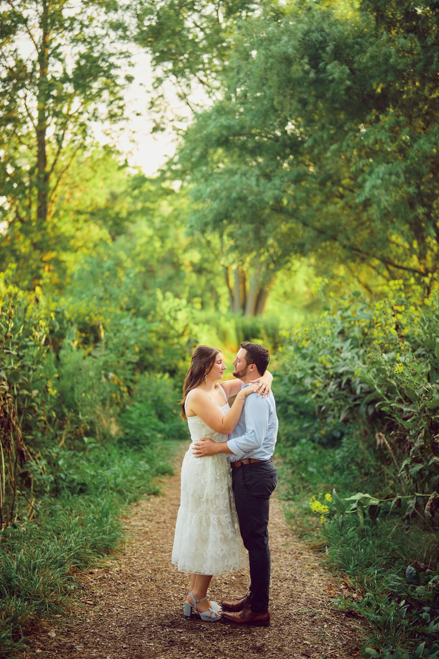 A romantic couple embraces on a sunlit forest path surrounded by lush green foliage and trees.