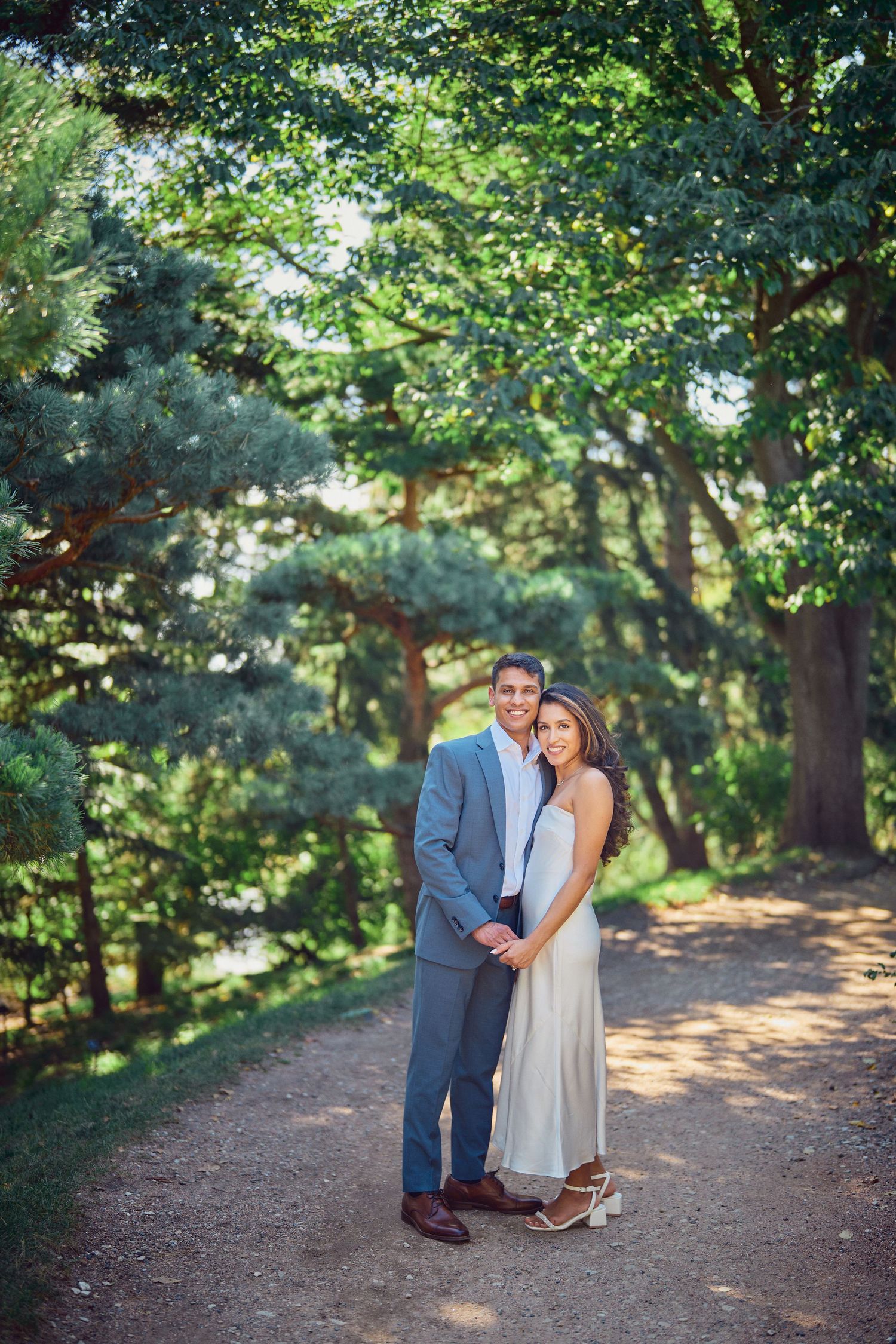 A couple stands together on a sunlit forest path surrounded by lush green trees and dappled light.
