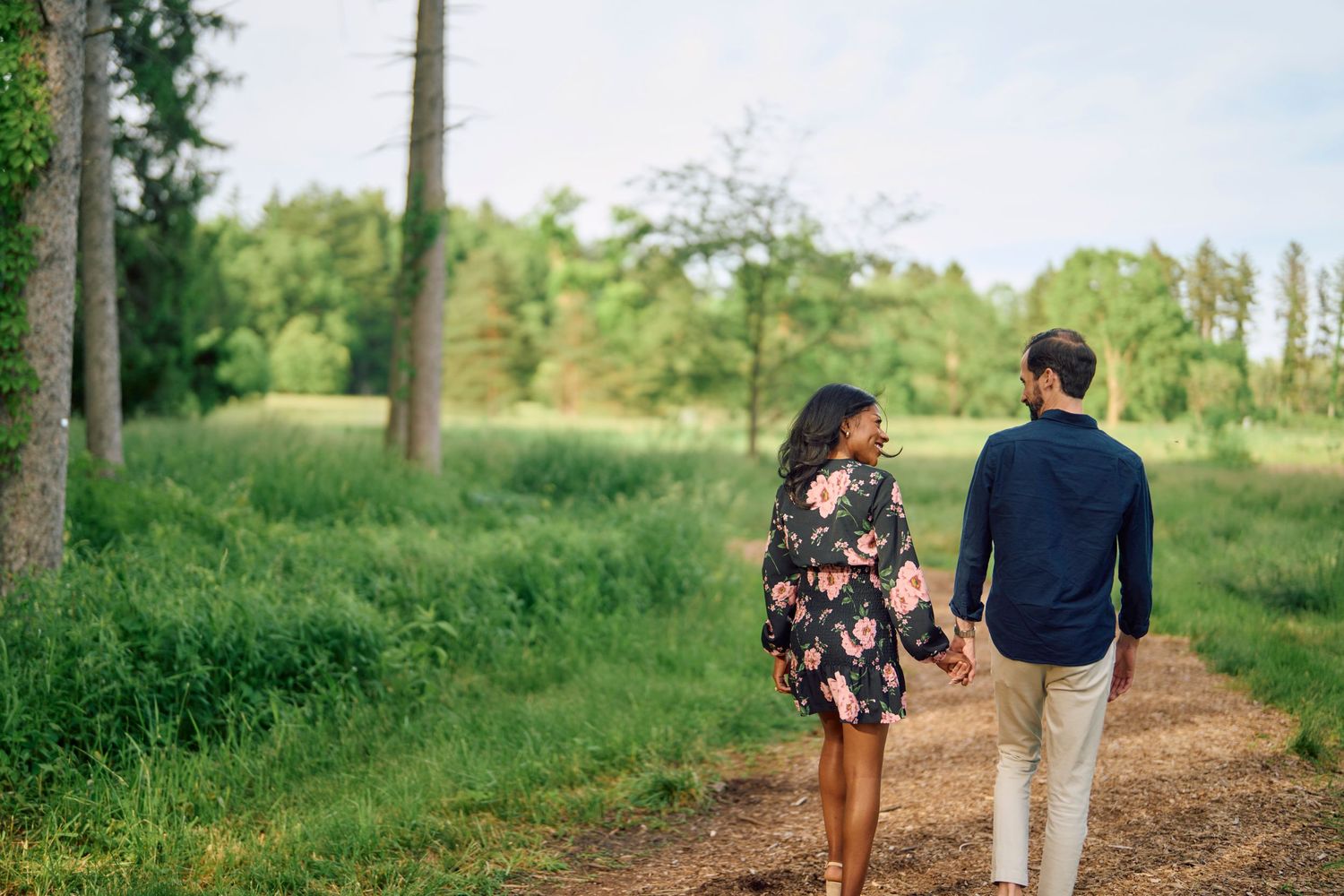 Two people in casual attire walk away from camera along a serene dirt path through lush green woodland on a sunny day.