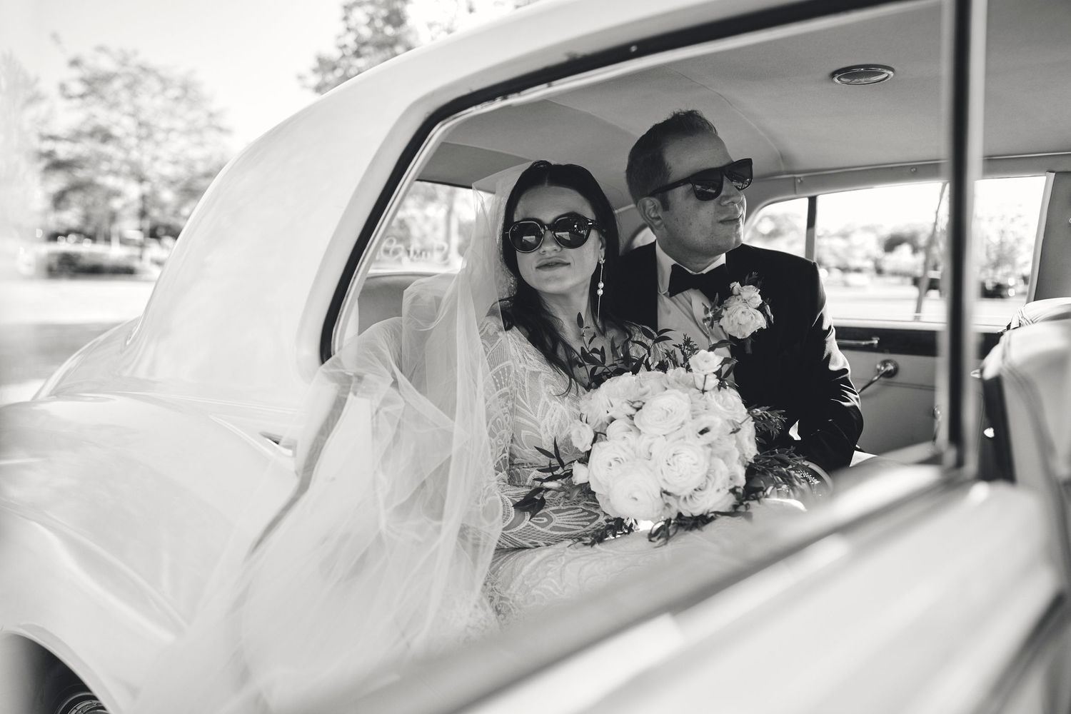 A vintage black and white wedding photo showing a couple sitting in a classic car with white flowers.