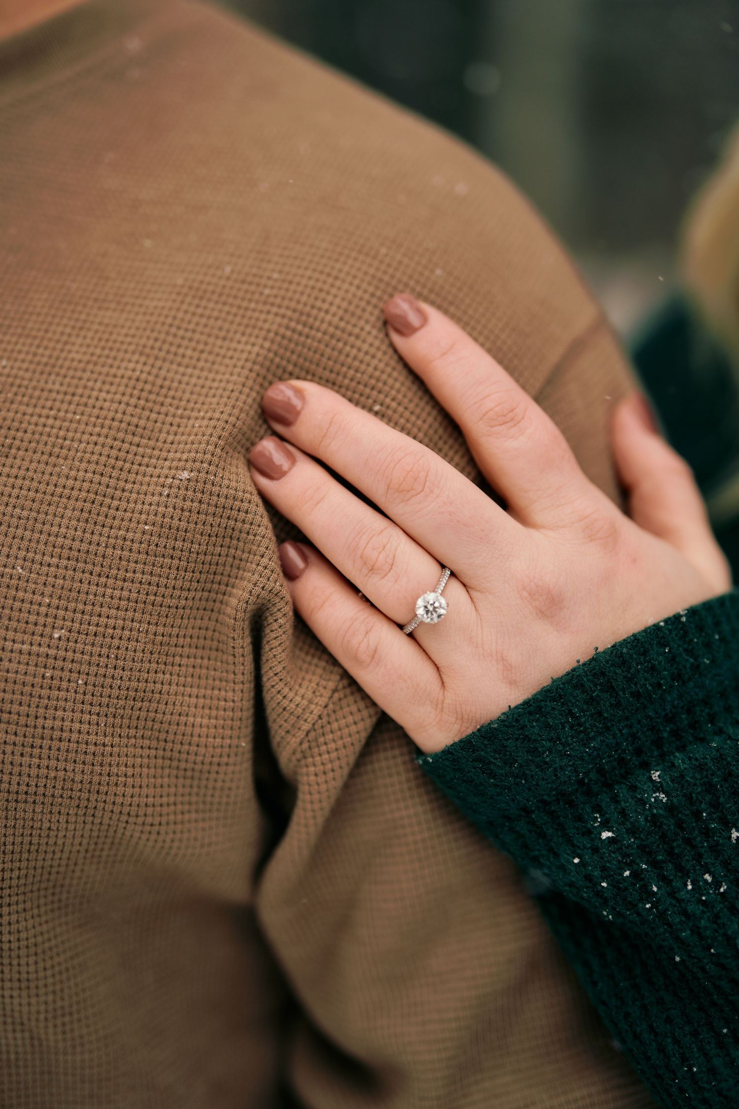 A hand wearing a sparkling diamond engagement ring rests against a brown fabric background.