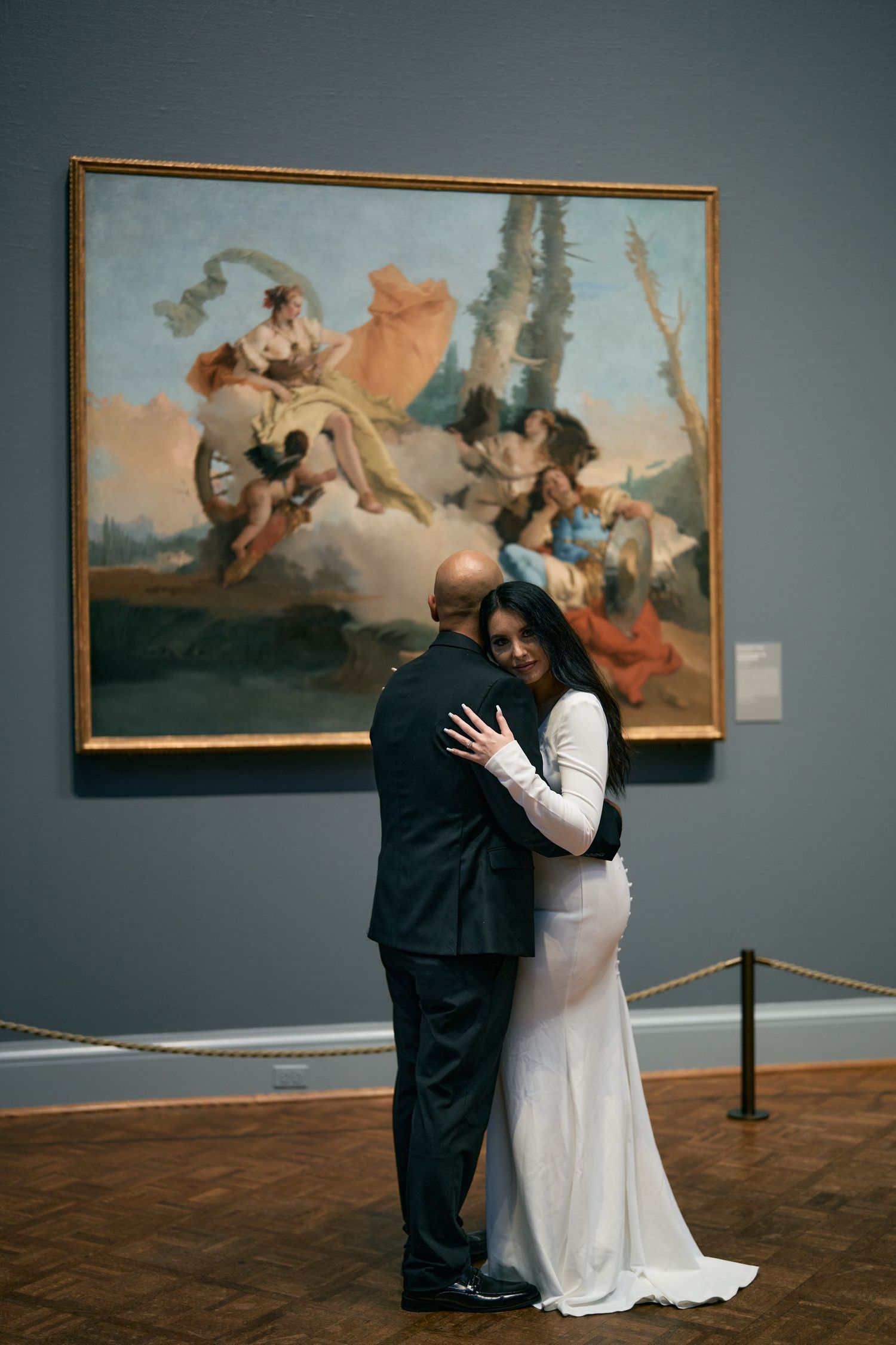 A couple embraces in front of a dramatic baroque painting in an art museum with wooden floors and gray walls.