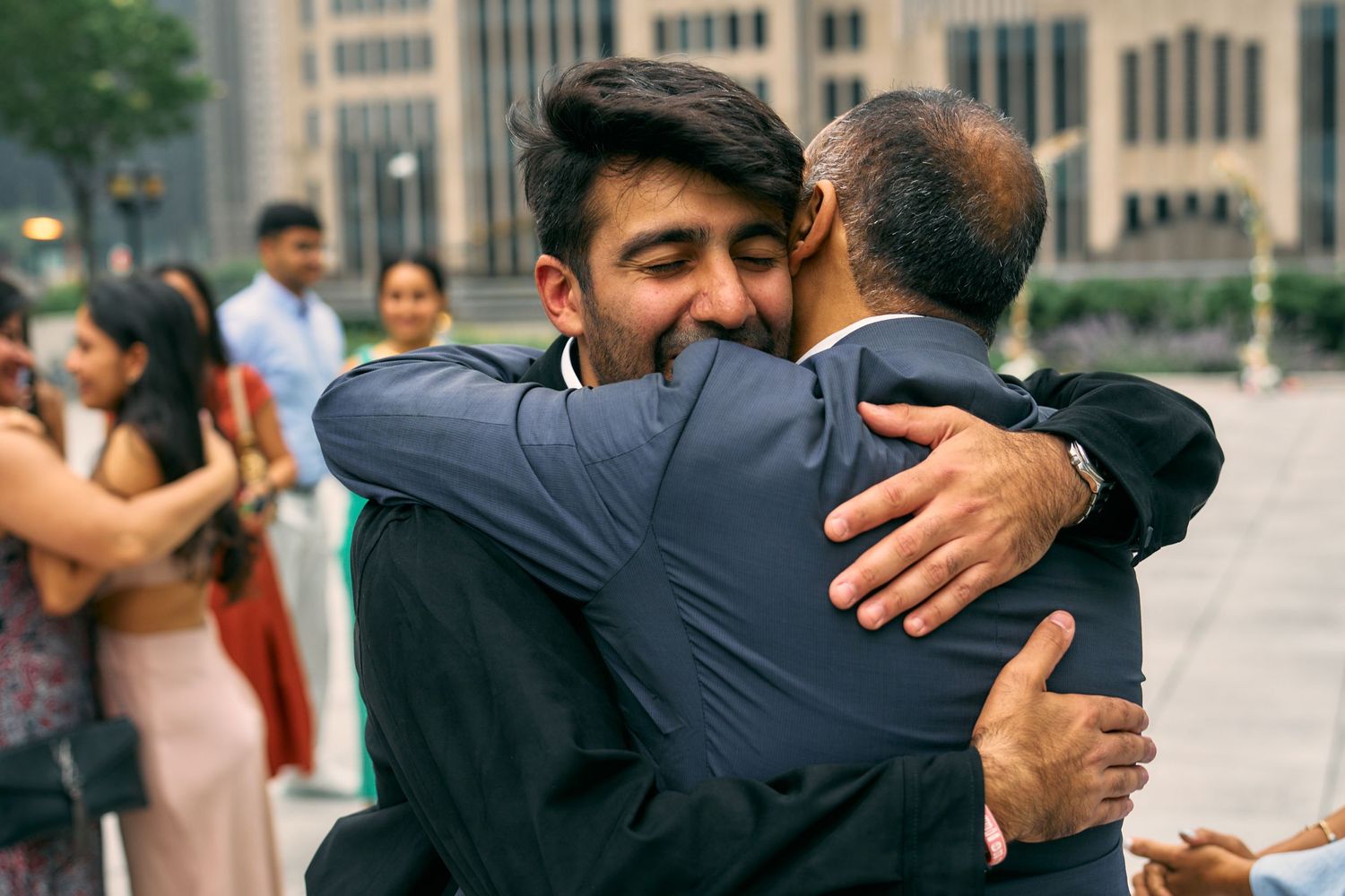Two people share a warm embrace outdoors with city buildings and other people visible in the background.